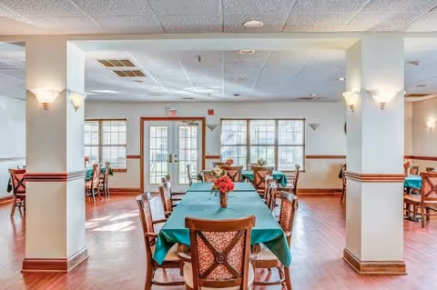 A bright and spacious dining room with wooden floors and multiple tables covered with green tablecloths. Each table is surrounded by wooden chairs with patterned cushions, and small flower arrangements are placed on the tables. Large windows allow natural light to fill the room, and there are wall sconces providing additional lighting.