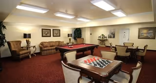 A cozy common room with a red carpet, featuring a pool table in the center, a checkers board on a wooden table in the foreground, several upholstered chairs and sofas, framed artwork on the walls, and soft overhead lighting.