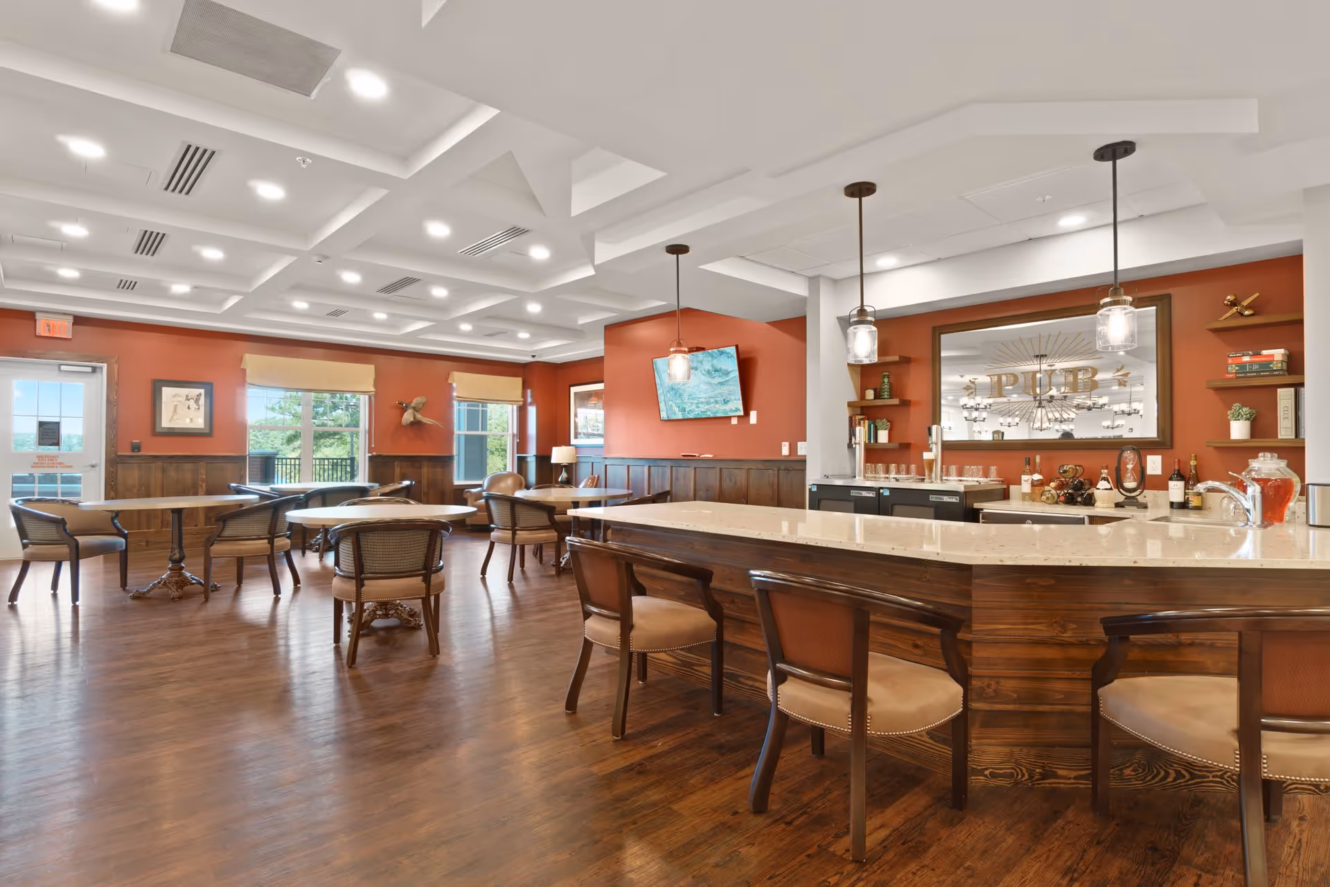 A spacious senior living common area with wooden floors, multiple round tables with chairs, and a large bar counter with stools. The walls are painted a warm reddish color with wood paneling on the lower half. Pendant lights hang above the bar, and a large mirror with the word 'PUB' is mounted on the wall behind the bar. There are windows letting in natural light and framed artwork on the walls.