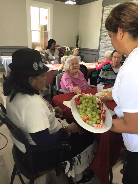 A group of elderly women seated around a table in a communal dining area. A caregiver is serving a platter of green grapes and other snacks to the women. The room has light-colored walls, a window, and a plant in the corner.