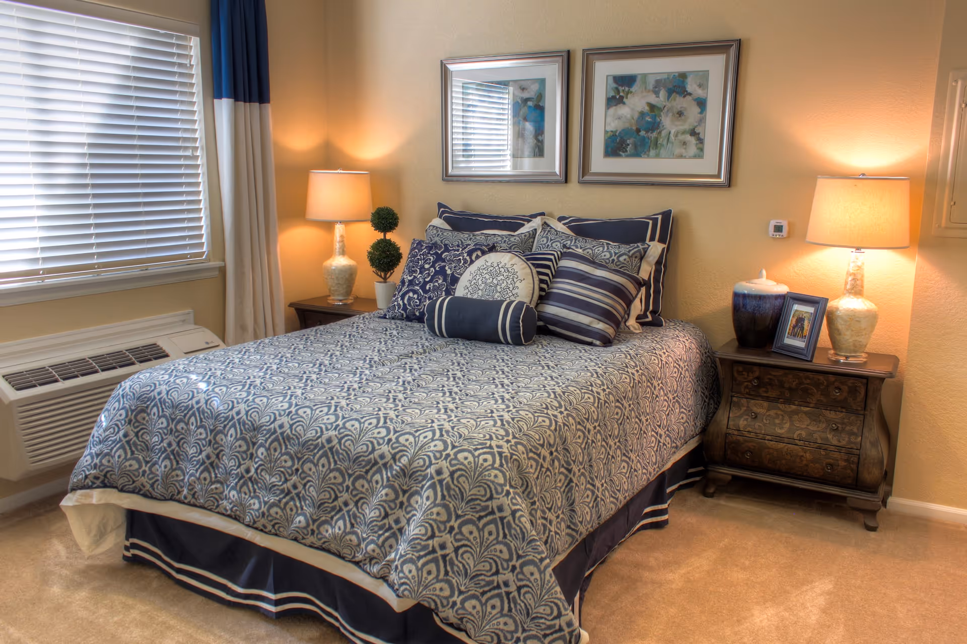 A neatly made bed with a blue and white patterned bedspread and multiple decorative pillows in a bedroom. There are two framed floral artworks hanging on the wall above the bed. On each side of the bed, there are wooden nightstands with lamps, a small plant, a framed photo, and a decorative jar. A window with closed blinds and curtains is visible on the left side of the room.