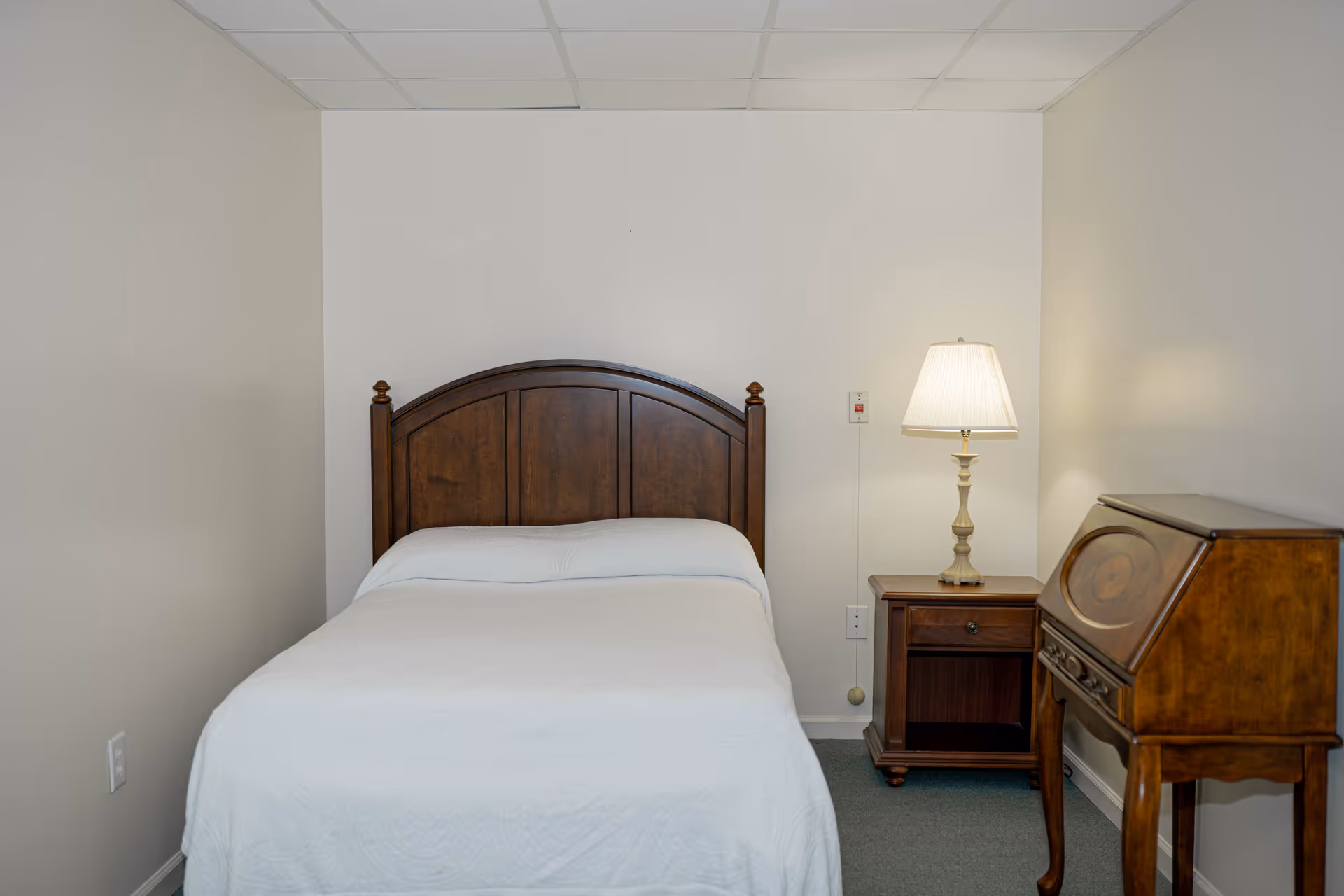 A simple bedroom with a wooden headboard bed covered with a white quilt, a wooden nightstand with a lamp, and a wooden writing desk against a plain white wall.