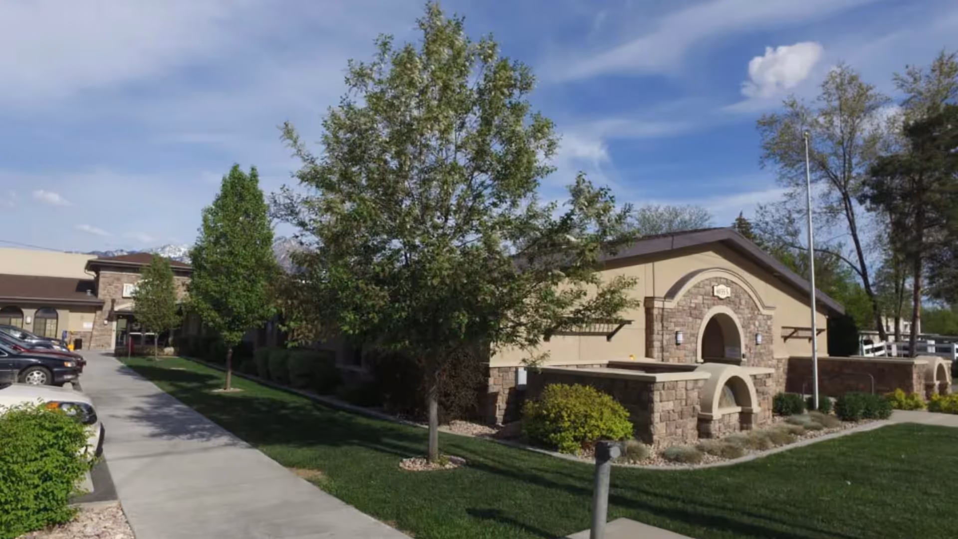 Exterior view of Paramount Health and Rehabilitation facility showing a stone and stucco building with arched entryways, surrounded by green grass, trees, and a sidewalk leading to the entrance. Several parked cars are visible on the left side under a partly cloudy sky.
