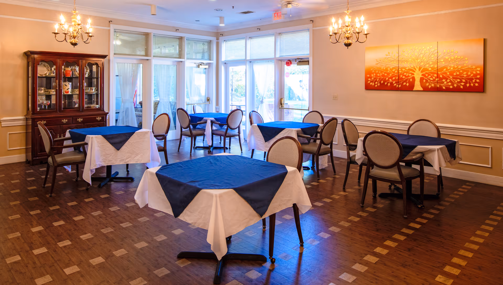 Dining room with several tables covered in white and navy tablecloths, upholstered chairs, chandeliers, a china cabinet, and large glass doors/windows.