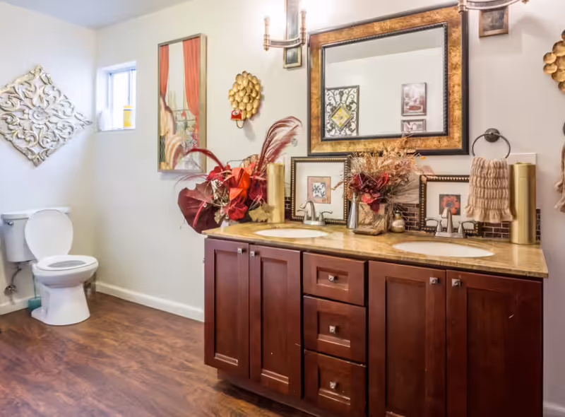A bathroom with a double sink vanity featuring dark wood cabinets and a beige countertop. Above the sinks is a large framed mirror with two wall-mounted light fixtures. The countertop is decorated with floral arrangements and framed pictures. To the left, there is a white toilet next to a small window. The walls are adorned with decorative wall art, and the floor has a wood-like finish.