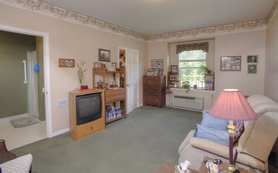 A cozy living room in a senior living facility with a beige couch, a side table with a lamp, a wooden TV stand with an old-style television, a wooden shelving unit, and a window with a valance. The room has green carpet and floral wallpaper border near the ceiling. An open door leads to a bathroom with a shower visible.