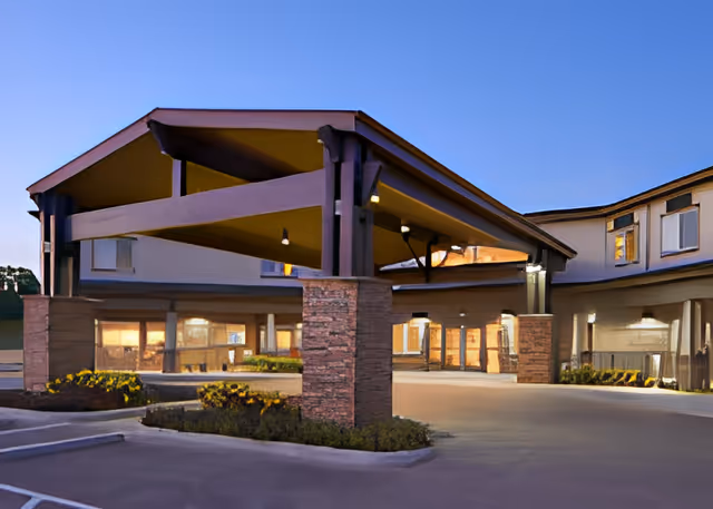 Exterior view of the entrance to Ralston Creek Neighborhood Assisted Living & Memory Care facility during dusk, featuring a covered drop-off area with stone pillars and warm interior lighting visible through large windows.
