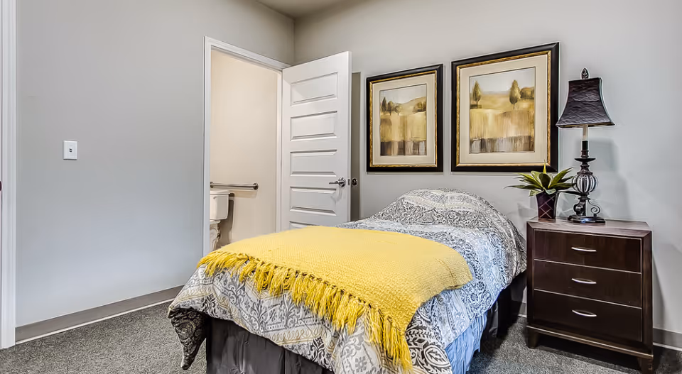 A small bedroom with a single bed covered in a patterned bedspread and a yellow throw blanket. Next to the bed is a dark wooden nightstand with a lamp and a potted plant. Two framed landscape paintings hang on the wall above the bed. An open door reveals a bathroom with a toilet and grab bar.