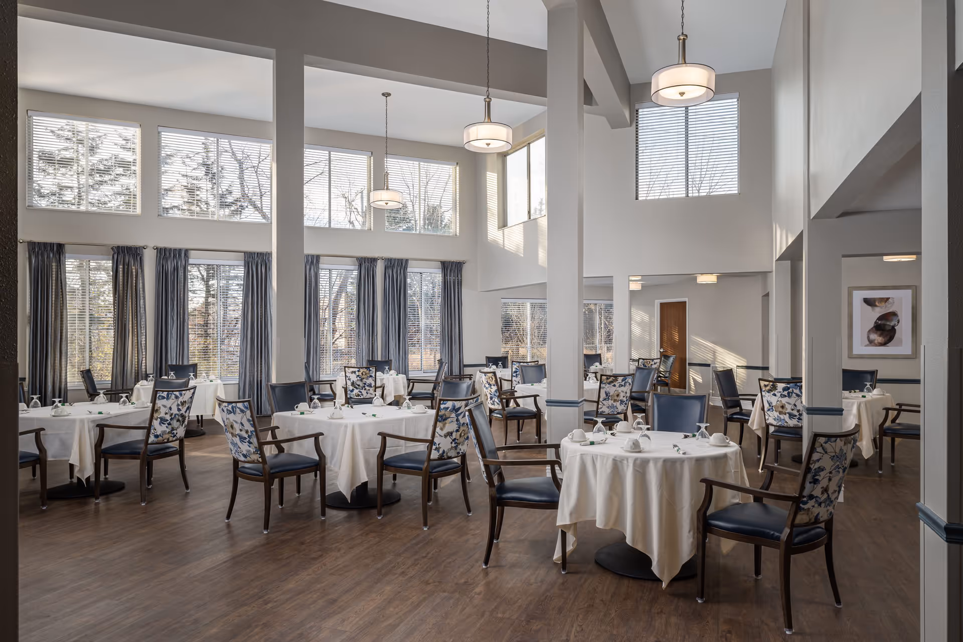 A bright and spacious dining room with multiple round tables covered with white tablecloths, each surrounded by chairs with floral and dark blue upholstery. Large windows with blinds and curtains allow natural light to fill the room. The room features high ceilings with hanging pendant lights and wooden flooring.