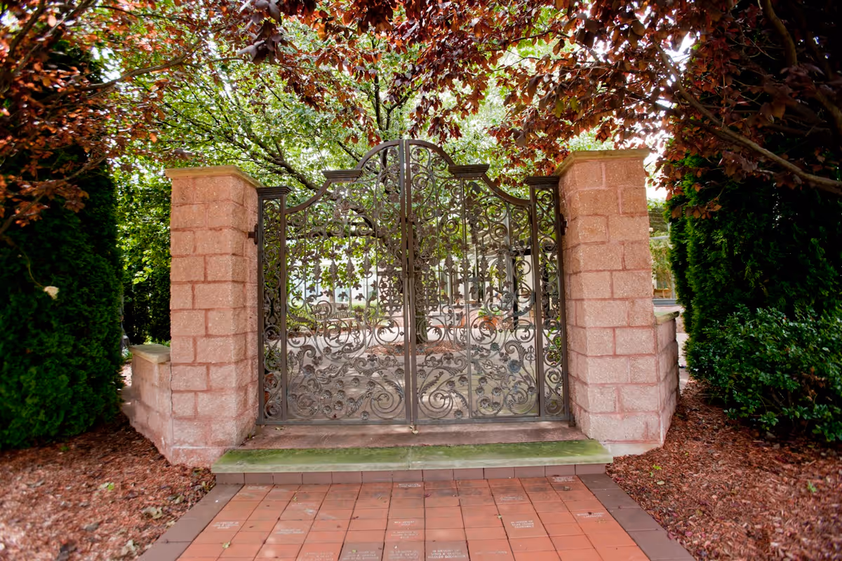 Ornate wrought iron gate set between two stone pillars with lush green trees and bushes surrounding the entrance, and a tiled pathway leading up to the gate.