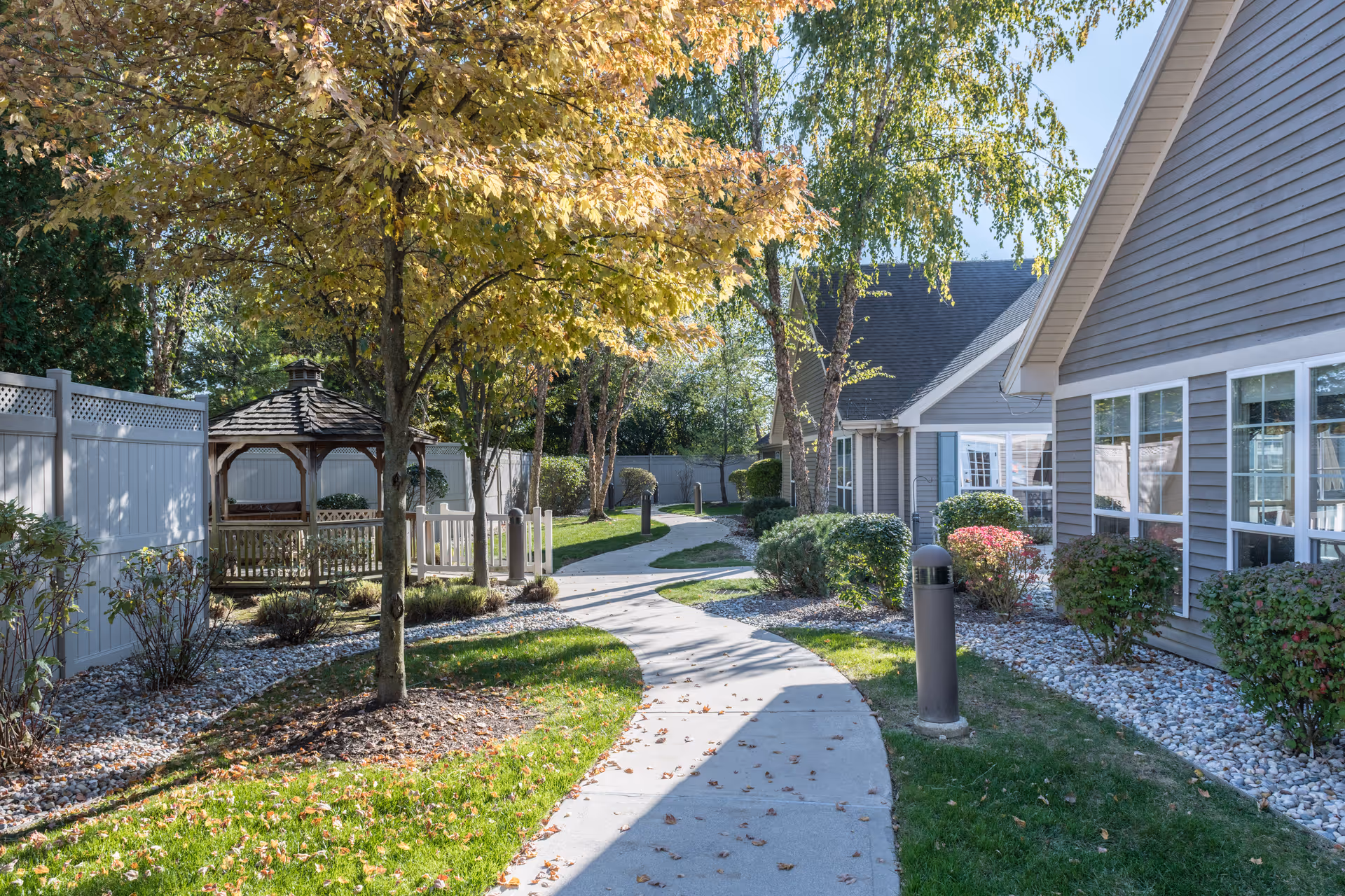 A winding concrete pathway through a landscaped garden area with green grass, bushes, and trees with autumn leaves. There is a wooden gazebo on the left side near a white fence, and a gray building with multiple windows on the right side. The sky is clear and blue.