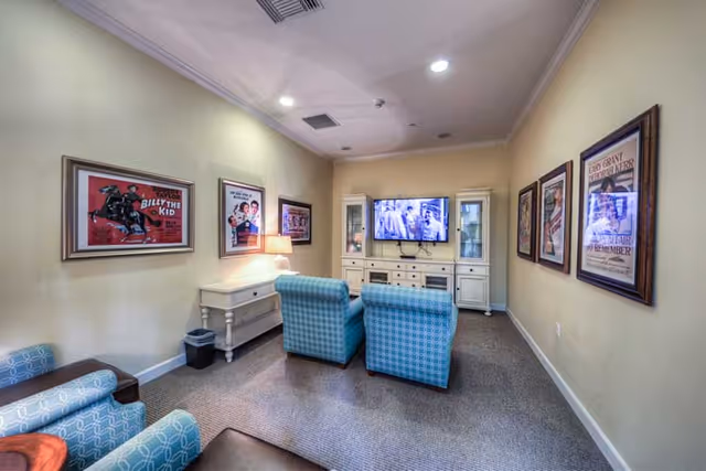 A cozy living room with two blue patterned armchairs facing a wall-mounted flat screen TV. The TV is flanked by two white display cabinets with glass doors. The walls are decorated with framed vintage movie posters. A small white desk with a lamp and a trash bin is positioned against the left wall. The room has beige walls and carpeted flooring.