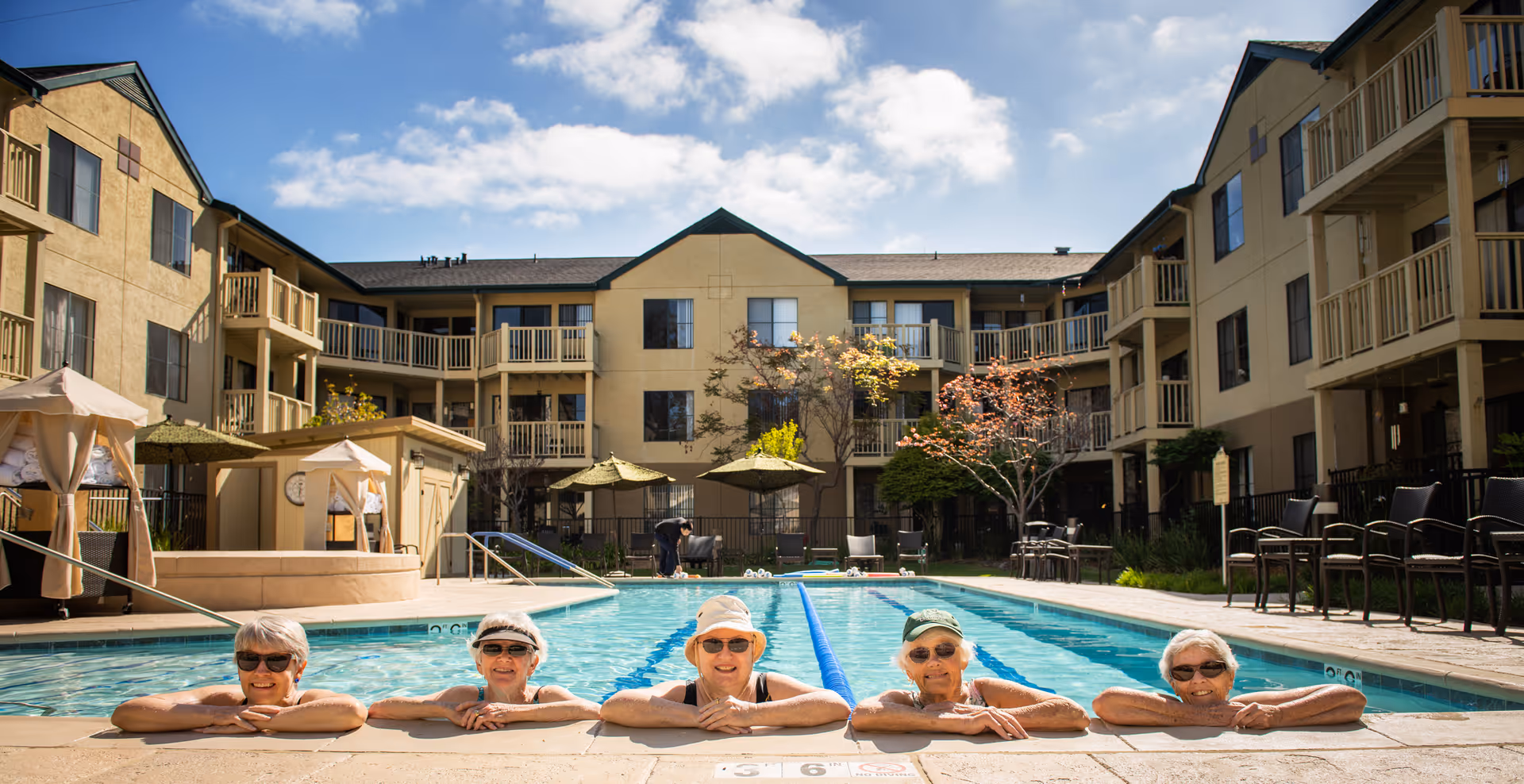 Five elderly women wearing sunglasses and swim caps are relaxing in an outdoor swimming pool at a senior living facility named Baywood Court. The pool is surrounded by a three-story building with balconies, patio umbrellas, and chairs under a partly cloudy sky.