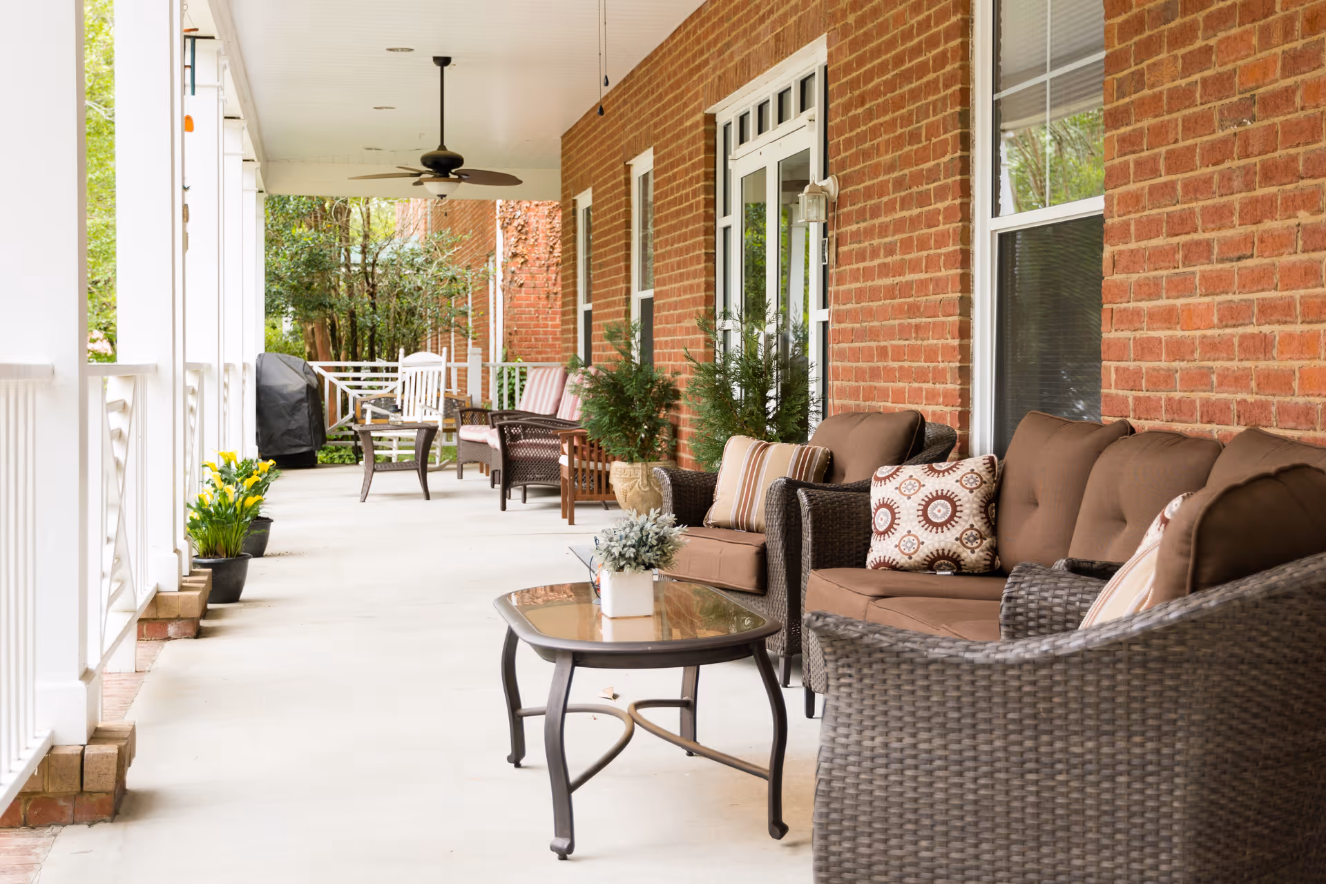 A spacious outdoor covered patio area with comfortable wicker seating including sofas and chairs with cushions and decorative pillows. There is a glass-top coffee table in the foreground, potted plants along the brick wall, ceiling fans overhead, and a grill in the background. The patio overlooks a green garden area.