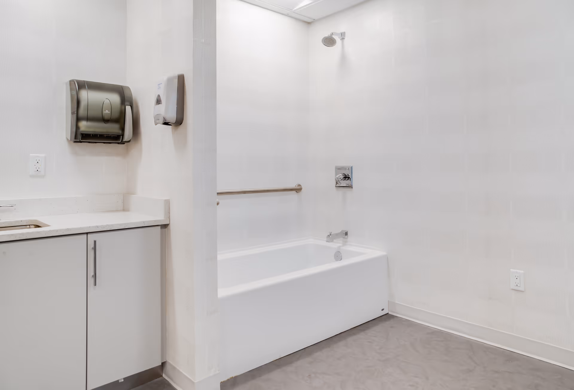 A clean, modern bathroom featuring a white bathtub with a wall-mounted showerhead and a grab bar. To the left, there is a countertop with a sink, a paper towel dispenser, and a soap dispenser mounted on the wall. The walls are light-colored with a subtle pattern, and the floor is a smooth gray surface.