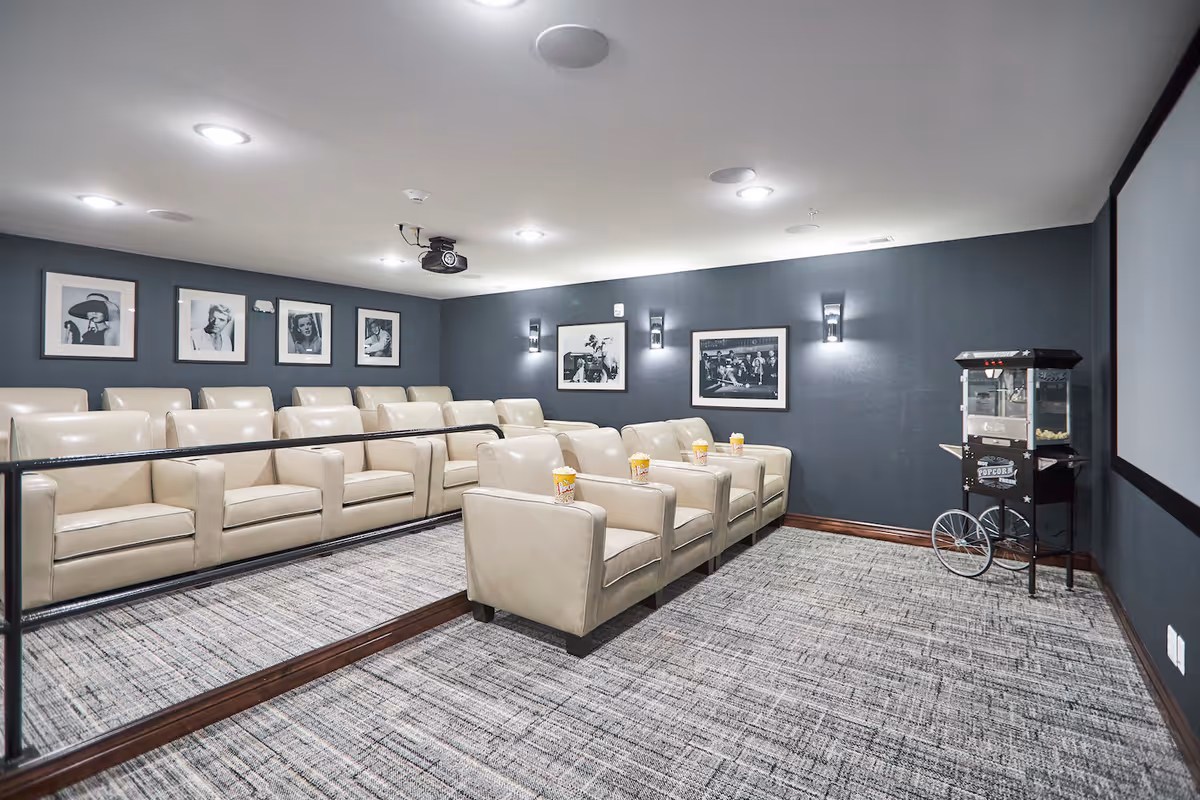 A small movie theater room with two rows of beige leather armchairs, each with a popcorn container on the armrest. The walls are dark gray and decorated with black and white framed photos of classic movie scenes and actors. A popcorn machine is positioned near the front screen, and ceiling lights illuminate the room.