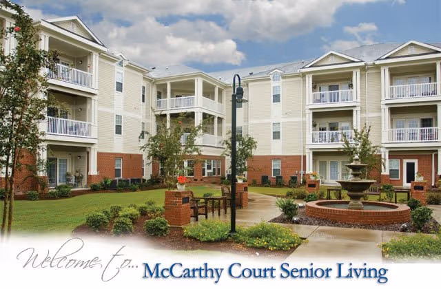 Exterior view of a senior living facility with three-story buildings featuring balconies, a landscaped courtyard with a fountain, benches, and a walking path under a partly cloudy sky.