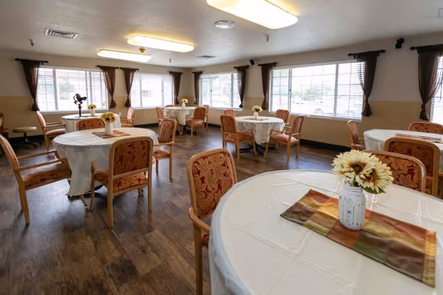 Bright dining room with round tables covered in white tablecloths, floral centerpieces, and upholstered chairs by large windows.