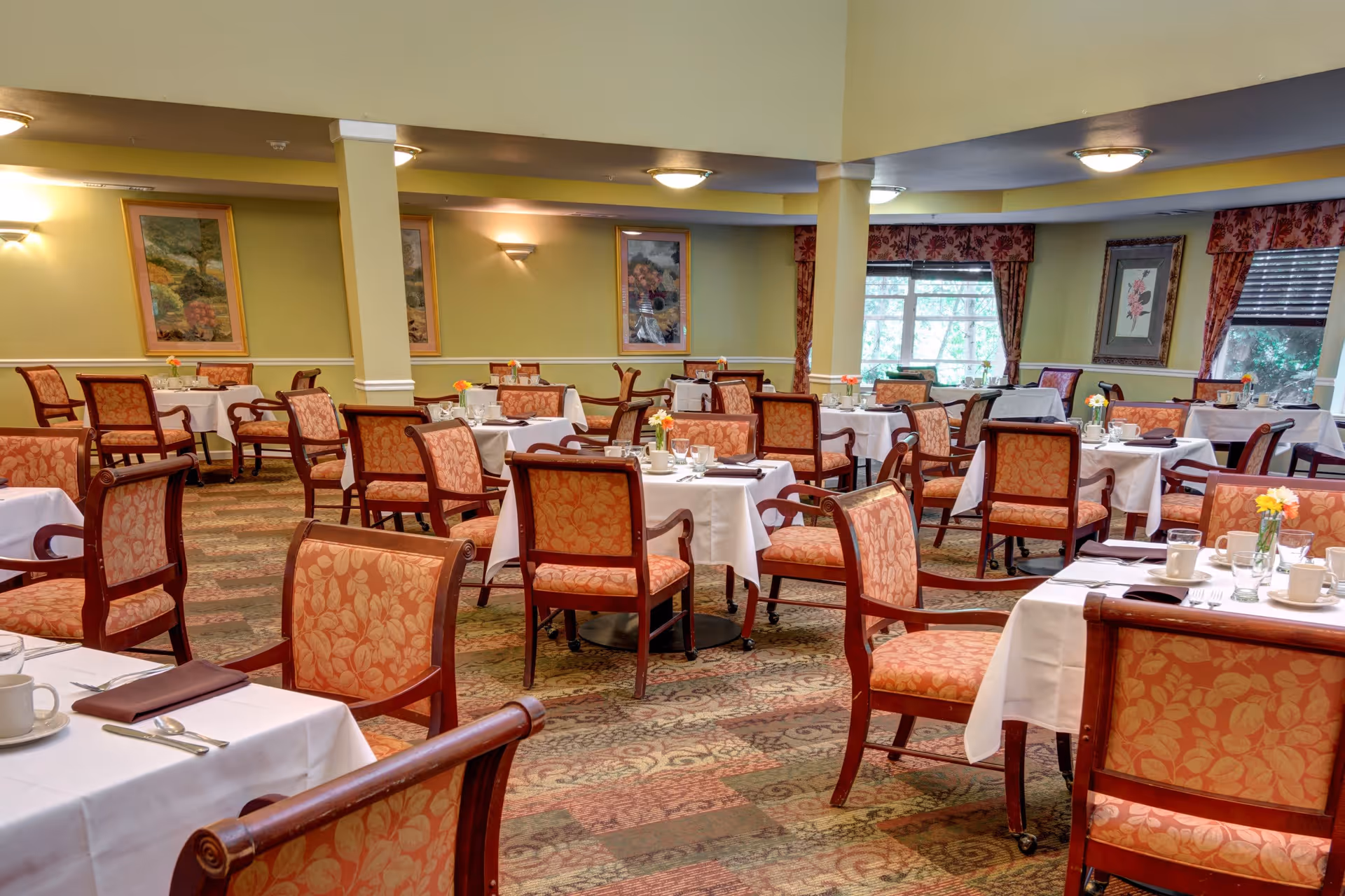 A dining room with multiple tables covered in white tablecloths, each set with cups, glasses, and silverware. The chairs have wooden frames with orange patterned upholstery. The room has soft yellow walls adorned with framed artwork and windows with floral curtains letting in natural light.