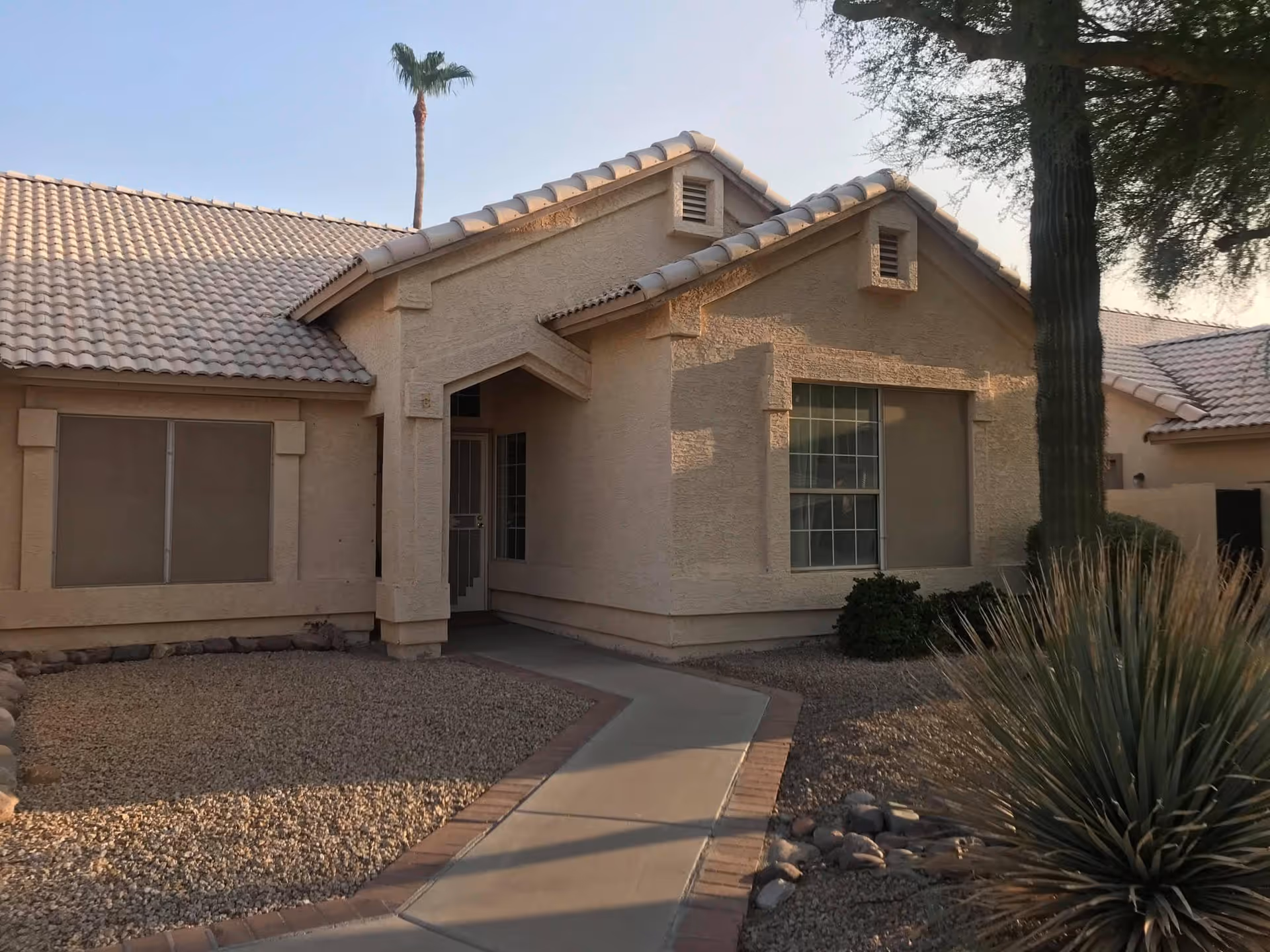 Front exterior of a single-story stucco house with a tiled roof, walkway, and desert landscaping including a cactus and palm tree.