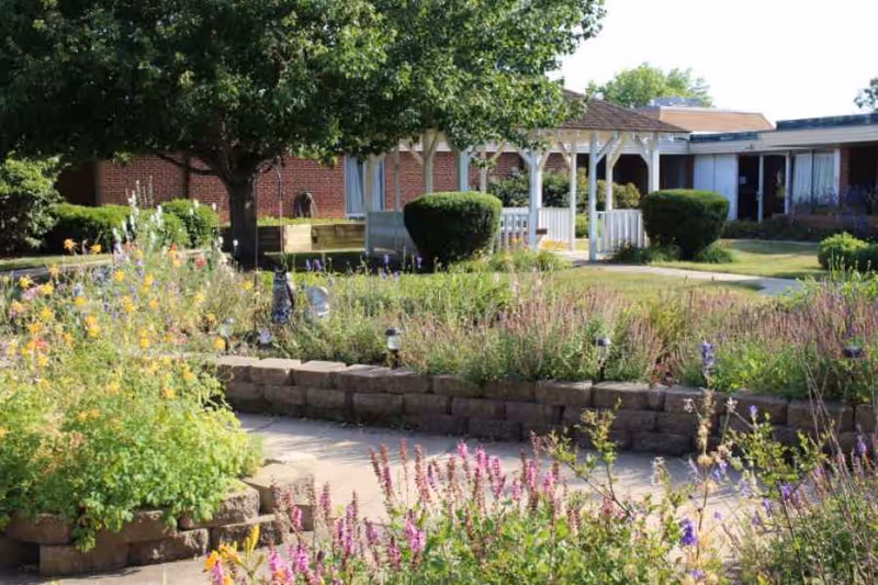 Sunny courtyard garden with flower beds, a white gazebo, trimmed shrubs, and a brick nursing home building in the background.