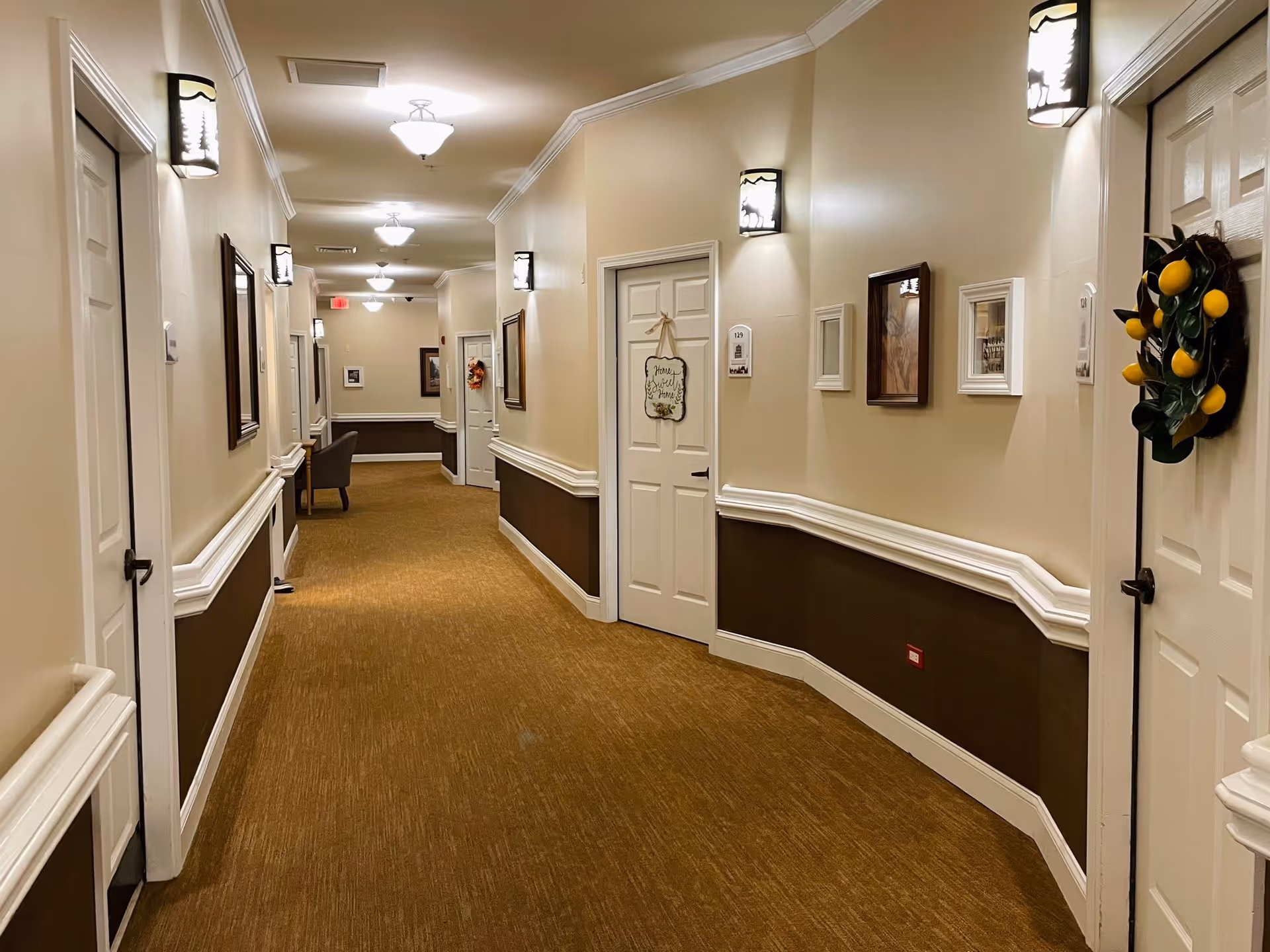 A well-lit indoor hallway in a senior living facility with beige walls and brown lower wall paneling. The hallway has multiple white doors, some decorated with wreaths and signs. Wall-mounted lights and framed pictures line the walls. A chair is visible at the far end of the hallway.