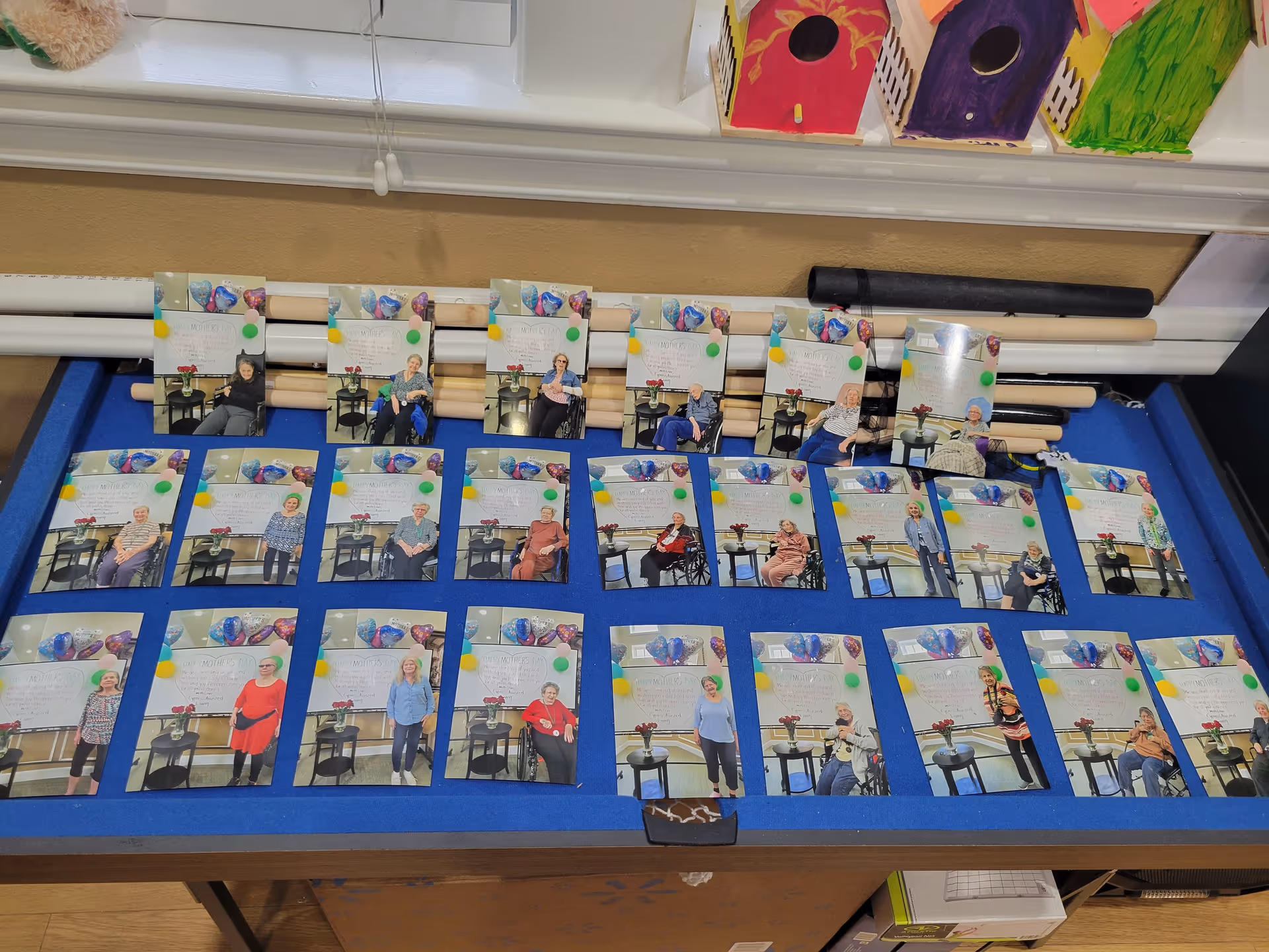 A display board with multiple photographs of elderly residents from Cypress Assisted Living - CyFair, each photo showing a resident posing next to a small table with a vase of flowers. The photos are arranged in rows on a blue felt board, with colorful birdhouse decorations visible on a shelf above.