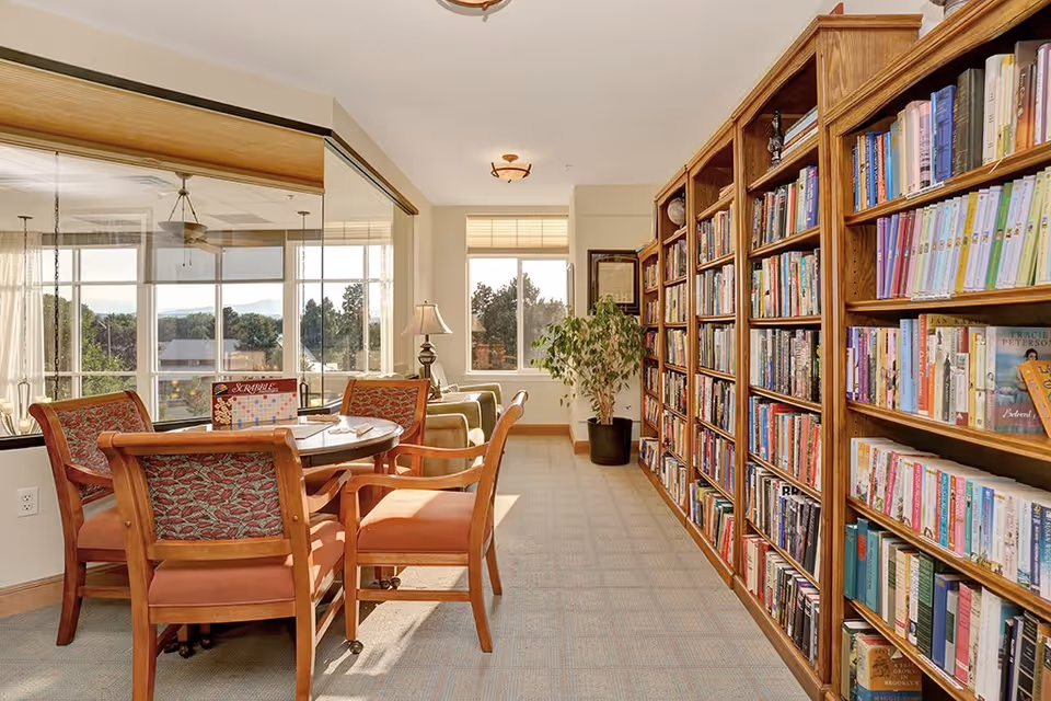 A bright and cozy library room with a large wooden bookshelf filled with books on the right side. In the center, there is a round table with four wooden chairs upholstered in orange fabric. On the table, there is a Scrabble game set up. The room has large windows letting in natural light and a view of trees outside. A green armchair and a floor lamp are visible near the windows.
