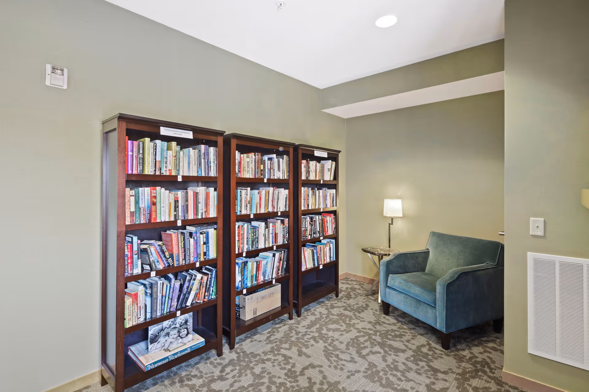 A cozy reading nook in a senior living facility featuring three wooden bookshelves filled with books, a teal upholstered armchair, a small side table, and a floor lamp against light green walls and patterned carpet.