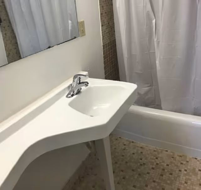 A bathroom with a white sink and faucet mounted on a white countertop. There is a large mirror above the sink, a bathtub with a white shower curtain, and mosaic tile flooring in shades of beige and brown.