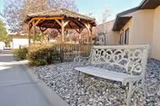 Outdoor area of a senior living facility featuring a decorative metal bench on a bed of rocks, a wooden gazebo surrounded by bushes, and a beige building wall in the background under a clear sky.