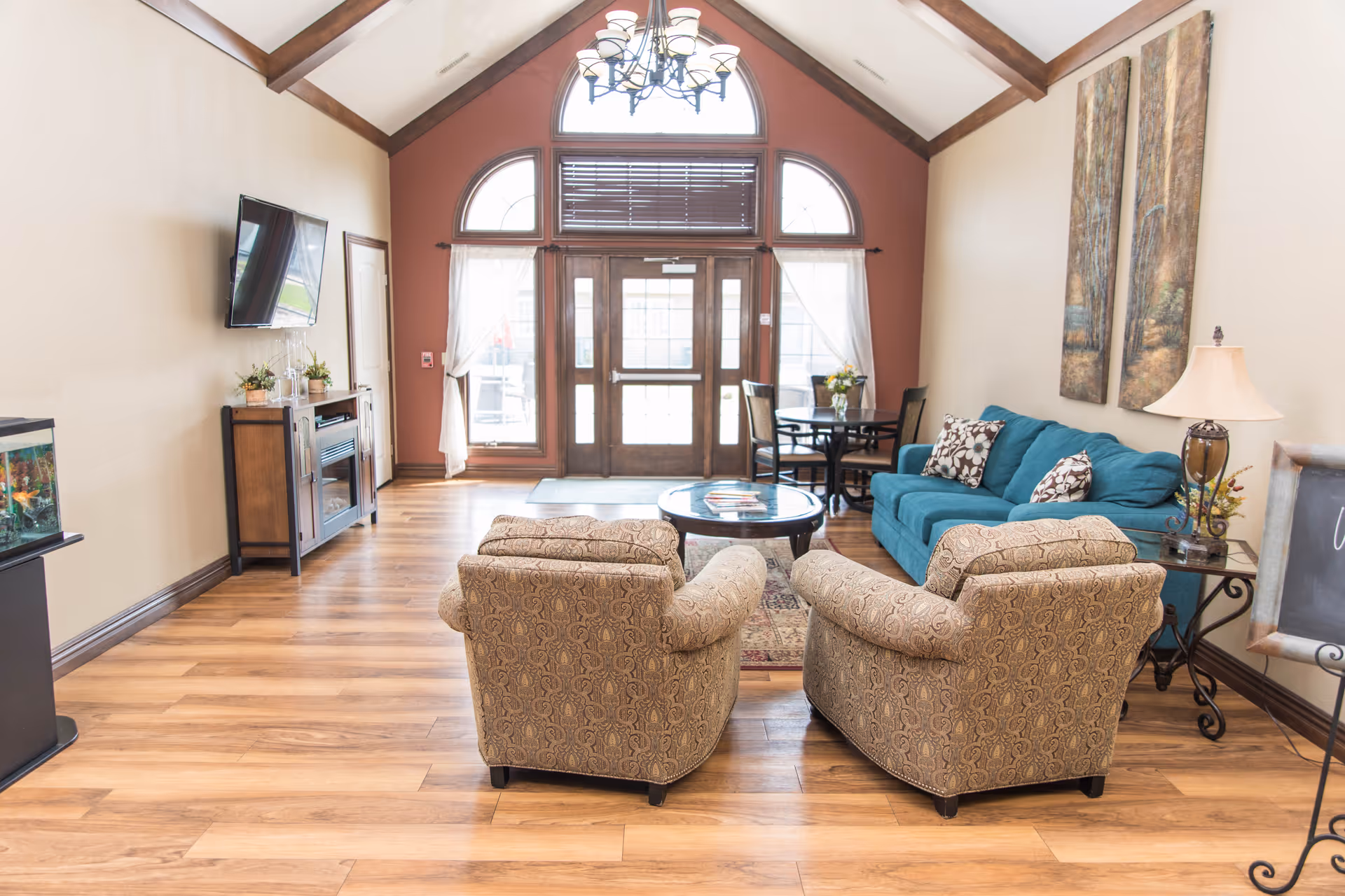 Bright communal living room with a teal sofa, two patterned armchairs, a round coffee table, a wall-mounted TV, and tall arched entryway windows.