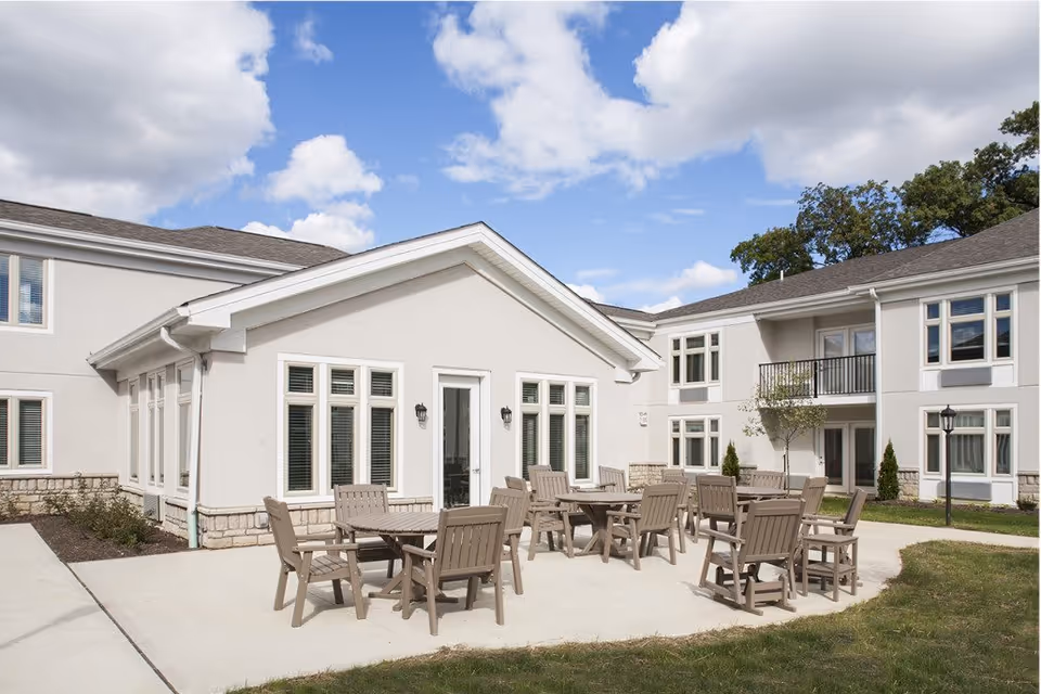 Outdoor patio area at The Ashford on Broad featuring several wooden tables and chairs on a concrete surface, surrounded by a two-story building with white walls and multiple windows under a partly cloudy blue sky.