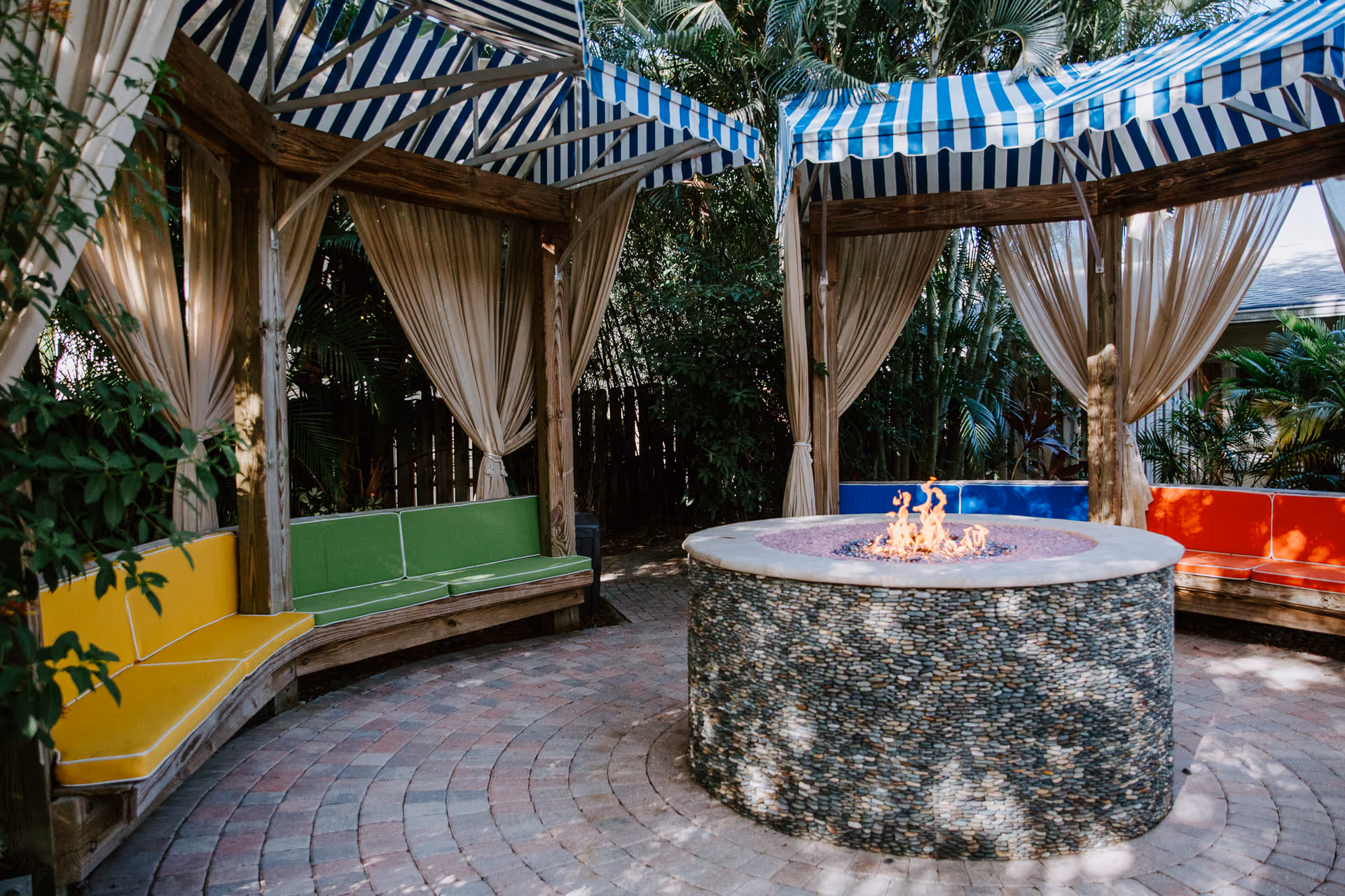 Outdoor courtyard with striped canopies, colorful cushioned benches, and a central fire pit.