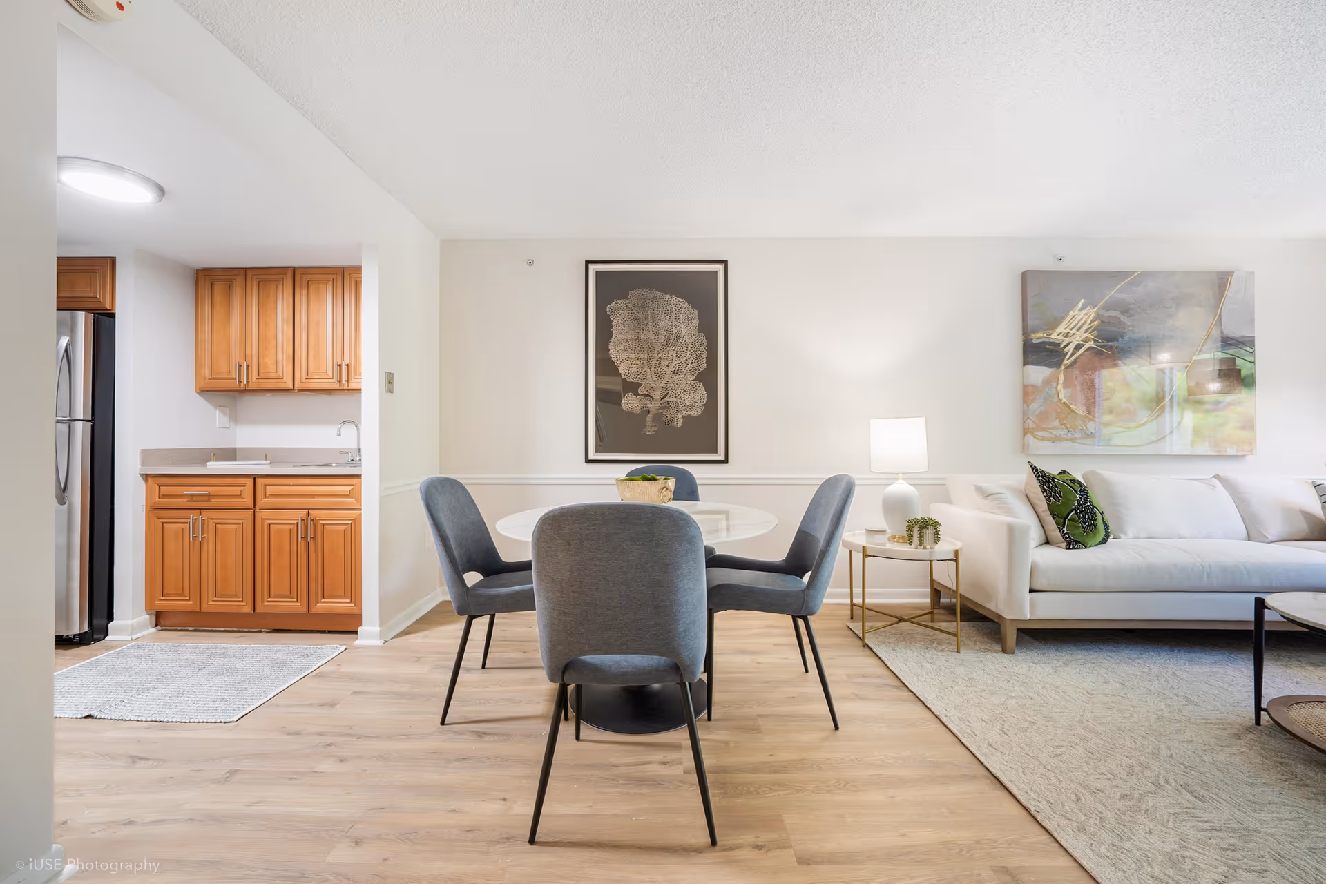 A bright and modern interior space showing a small dining area with a round white table and four gray upholstered chairs. To the left is a kitchen area with wooden cabinets and a stainless steel refrigerator. On the right side, there is a white sofa with green and patterned pillows, a small round side table with a white lamp, and abstract wall art above the sofa. The floor is light wood, and the walls are painted white.
