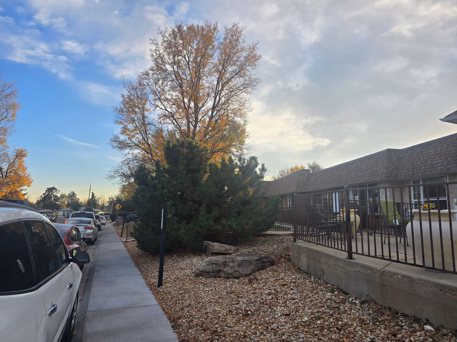 Sidewalk next to a parking area with several parked cars on the left and a low patio area with chairs and tables on the right, adjacent to a single-story building. Trees with autumn foliage are visible in the background under a partly cloudy sky.