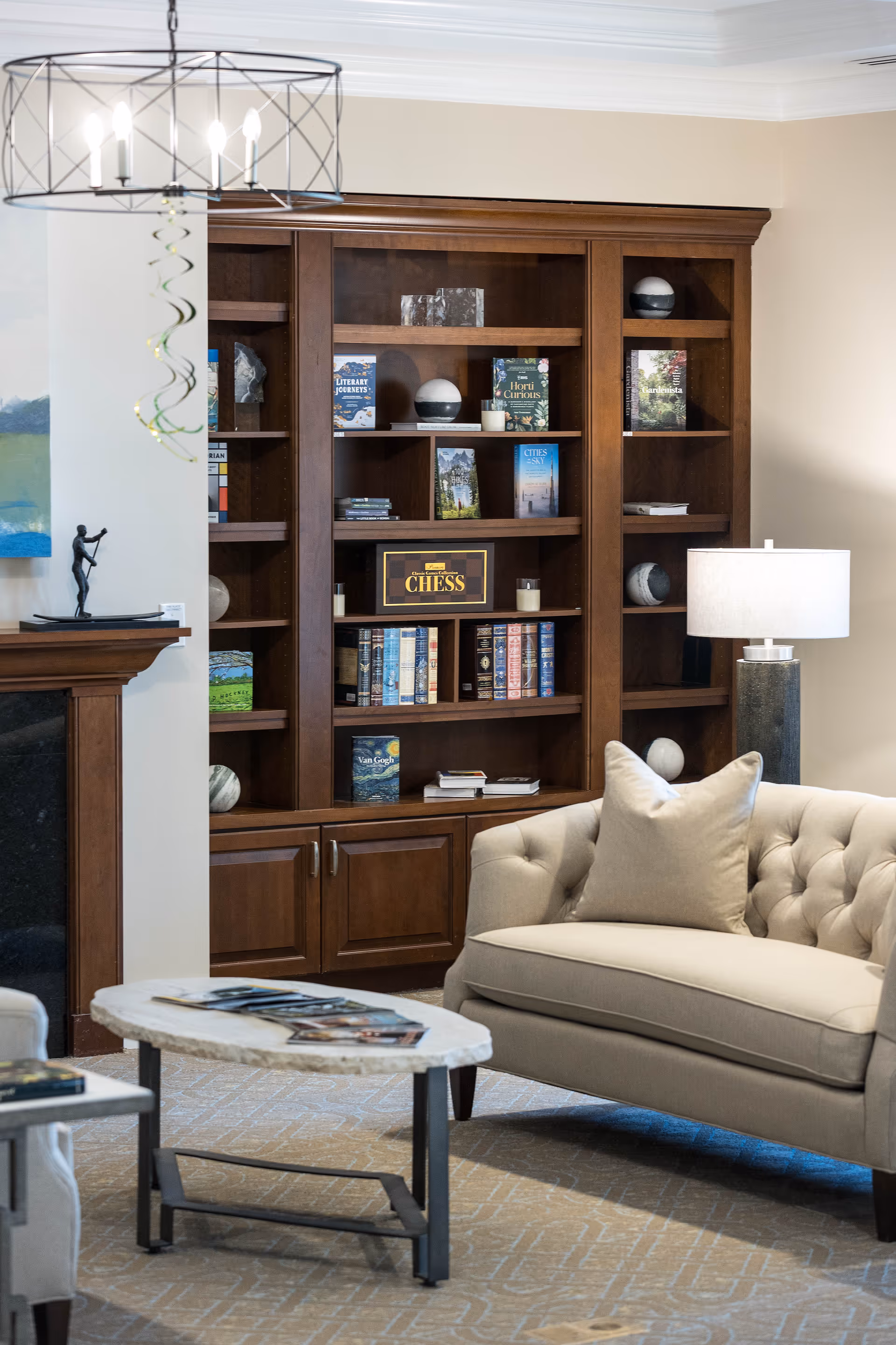 A cozy living room area featuring a beige tufted sofa with a matching pillow, a wooden bookshelf filled with books and decorative items, a modern table lamp on a side table, a round coffee table with magazines, and a fireplace mantel with a small sculpture. The room has a warm and inviting atmosphere with neutral tones and soft lighting.