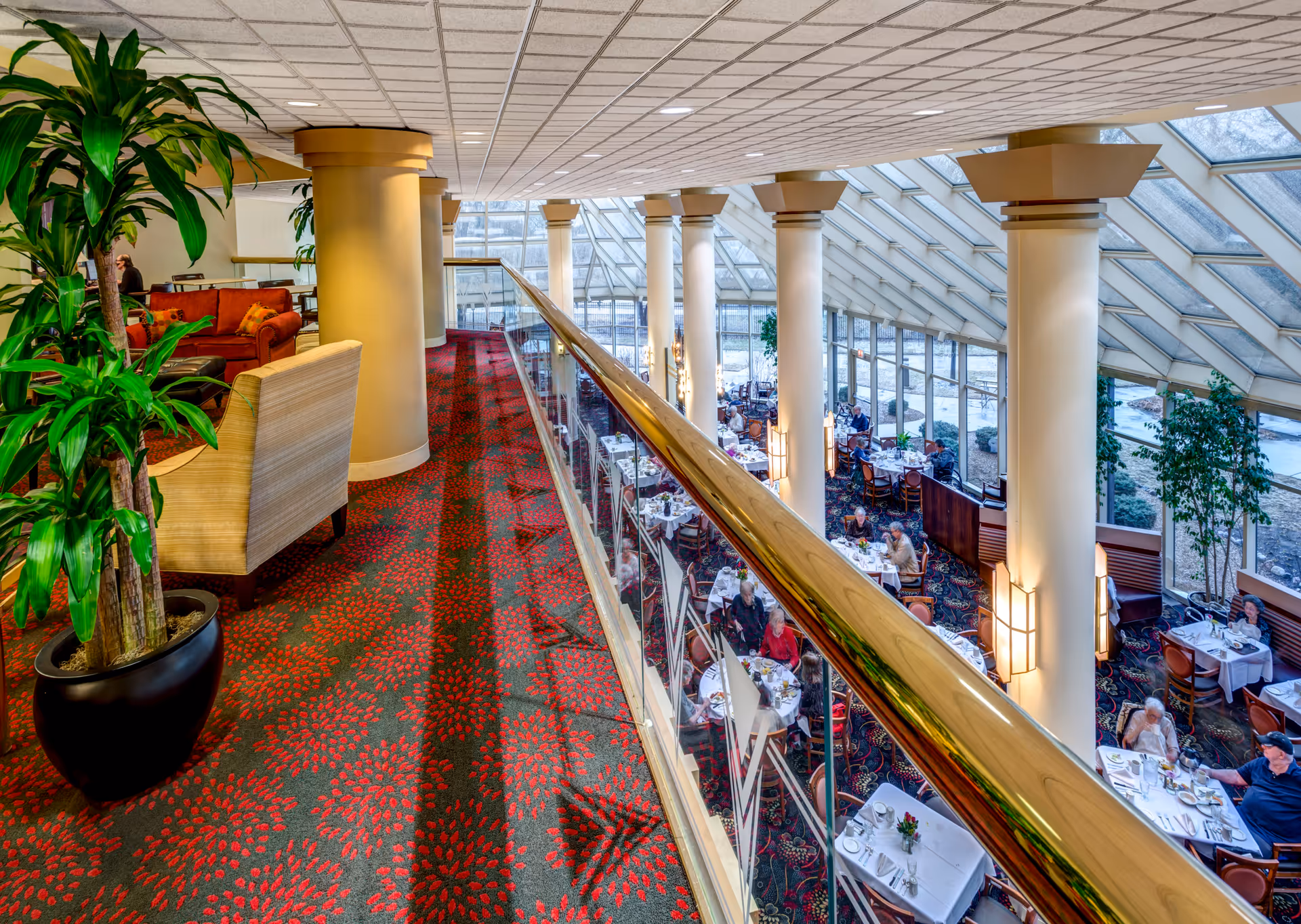 View from an upper level hallway overlooking a dining room with large windows and a glass ceiling. The hallway has a red patterned carpet, potted plants, and seating areas with chairs and sofas. The dining room below has tables set with white tablecloths and people seated, enjoying their meals.