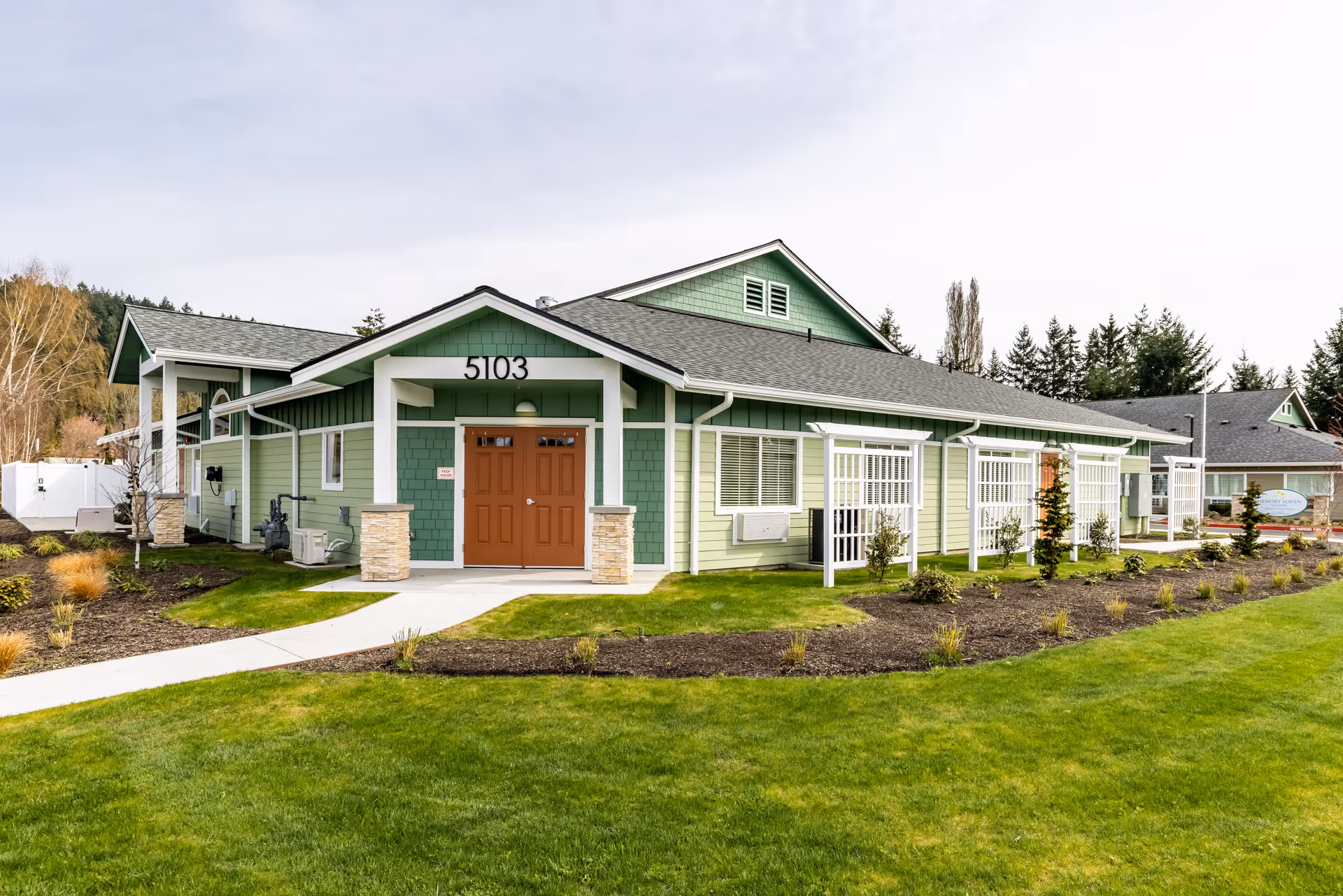 Exterior view of a single-story green building with white trim and a brown double door entrance, surrounded by a well-maintained lawn and landscaped garden beds. The building has the number 5103 above the entrance and is set against a backdrop of trees and a partly cloudy sky.