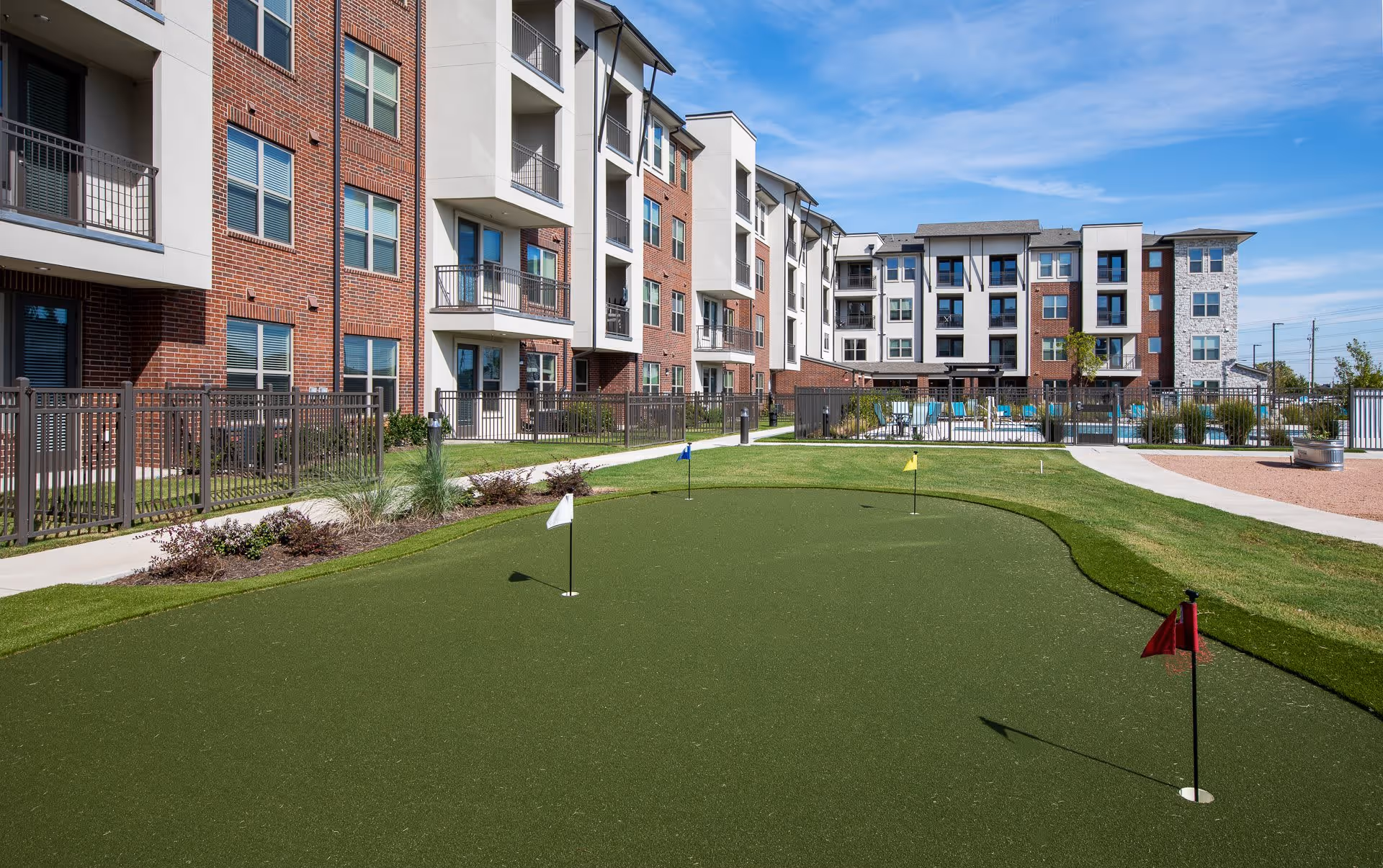 Outdoor putting green with four small flags in front of a multi-story residential building with balconies and windows under a blue sky.