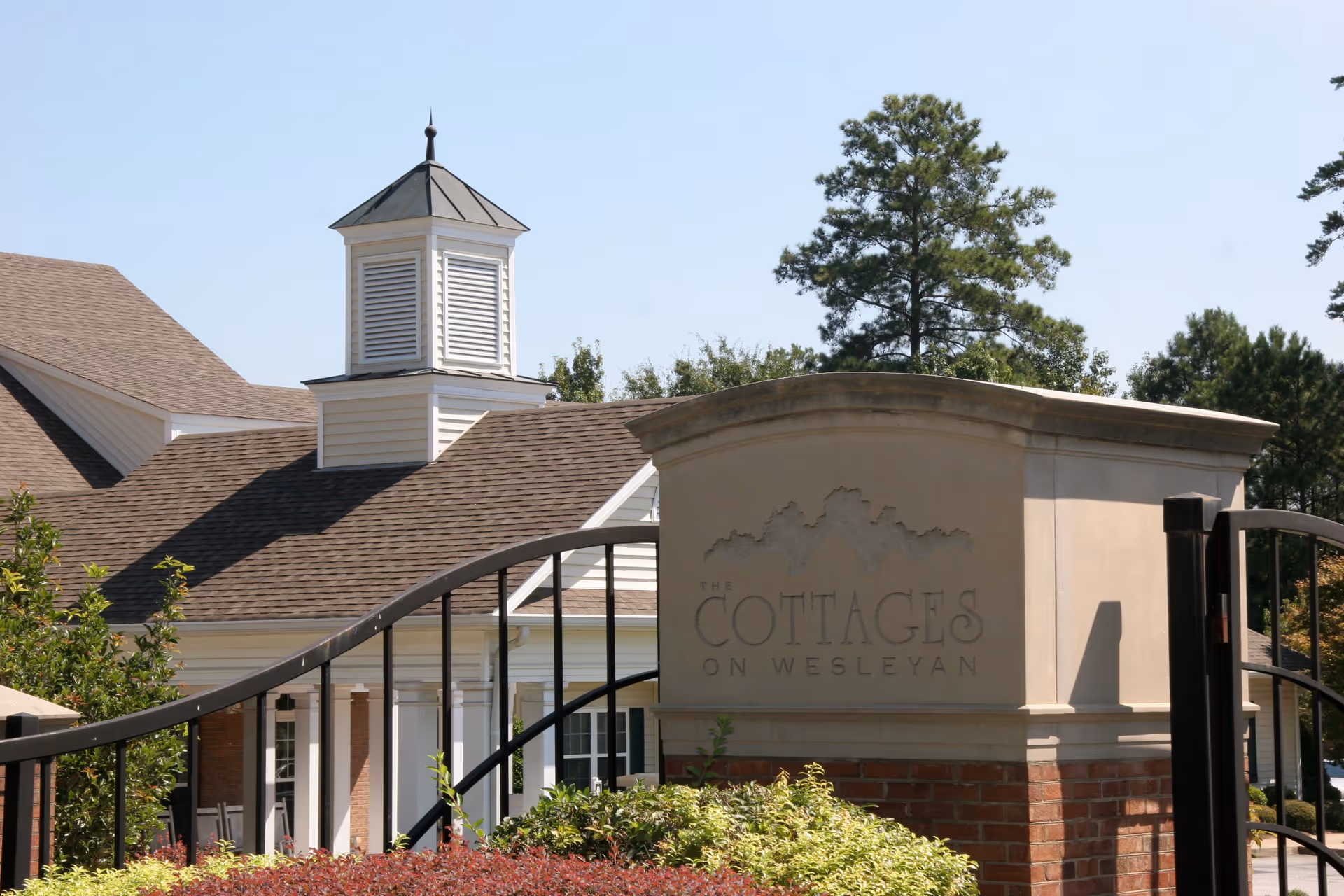 Entrance sign for The Cottages on Wesleyan with a gated fence and a building with a cupola in the background surrounded by trees and shrubs under a clear sky.
