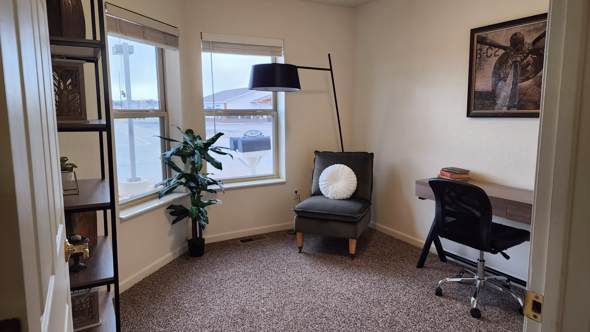 A small room with beige walls and carpeted floor featuring a gray cushioned chair with a round white pillow, a black floor lamp, a small desk with a black office chair, a few books on the desk, a tall plant near two windows, and a shelving unit on the left side.