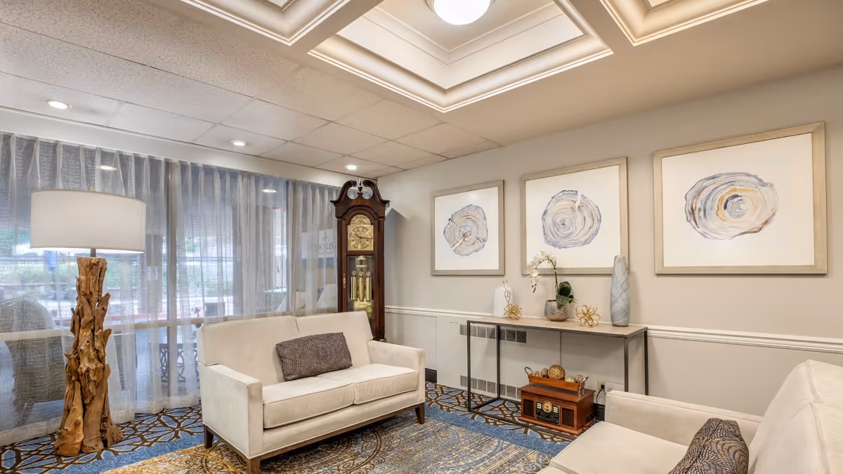 A cozy living room area with two beige sofas facing each other, each with a decorative pillow. Between them is a patterned carpet with blue and brown tones. A tall wooden floor lamp with a white shade stands to the left near sheer curtains covering large windows. A classic grandfather clock is positioned in the corner. On the right wall, there is a narrow console table decorated with vases and ornaments, beneath three framed abstract art pieces featuring circular designs in neutral colors. The ceiling has recessed lighting and a tray ceiling design.