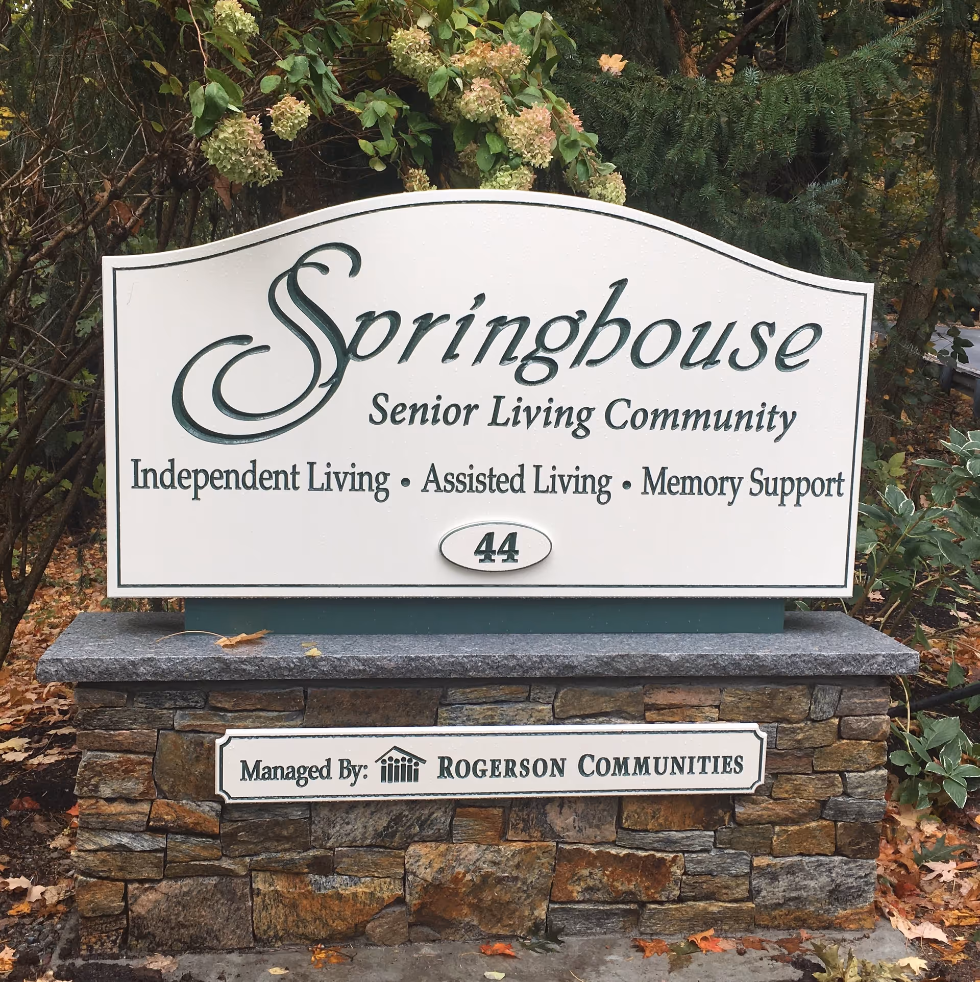 A stone-mounted outdoor sign reading "Springhouse Senior Living Community — Independent Living • Assisted Living • Memory Support" surrounded by plants and fallen leaves.