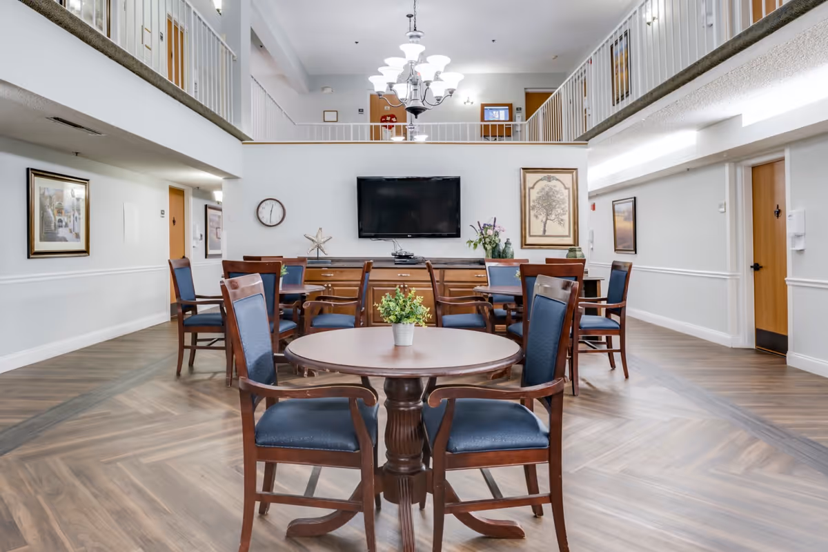 Interior view of a senior living facility common area with wooden round tables and chairs with blue cushions. The room has a high ceiling with a chandelier, a wall-mounted flat-screen TV, framed artwork, and a clock on the wall. There is a second-floor balcony overlooking the area.