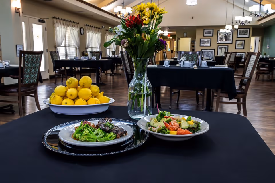 A dining room in an assisted living facility with tables covered in black tablecloths. In the foreground, a table displays a plate of cooked broccoli and meat, a bowl of fresh salad, a vase with colorful flowers, and a bowl filled with lemons. The room has wooden floors, chairs with patterned upholstery, and framed pictures on the walls.