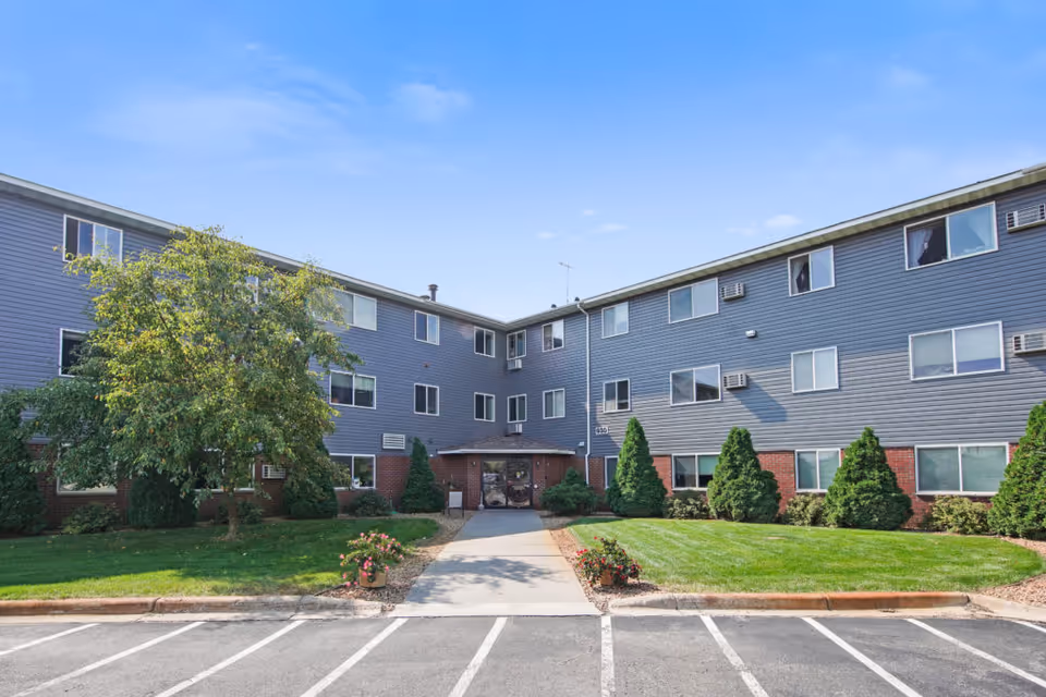 Exterior view of Keller Lake Commons, a three-story residential building with gray siding and red brick accents. The entrance is centered with a small covered porch, surrounded by green shrubs, trees, and a well-maintained lawn. There is a parking area in front with marked parking spaces under a clear blue sky.