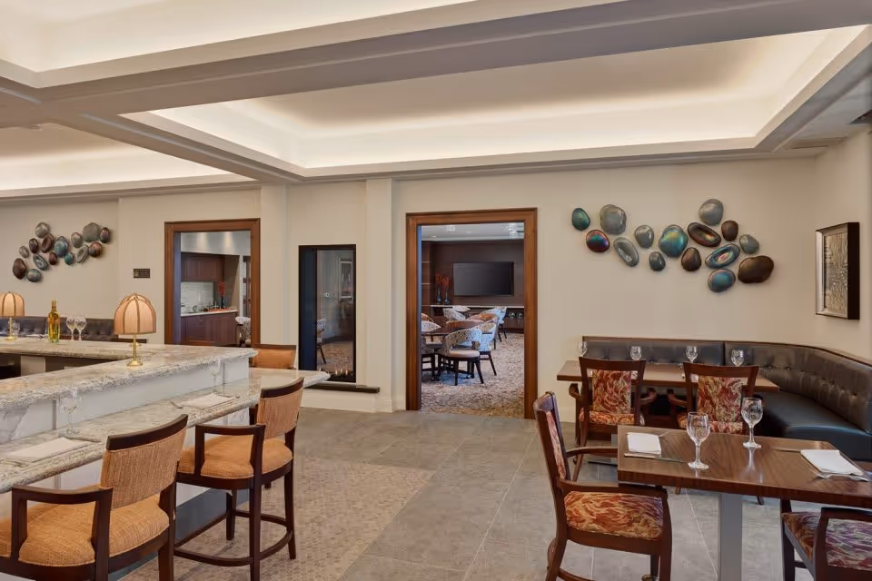 Interior view of a senior living facility dining area with a marble countertop bar and several bar stools on the left. To the right, there are wooden dining tables with chairs and a curved leather bench seat. The walls are decorated with modern art pieces. In the background, there is an open doorway leading to another room with additional seating and a large TV mounted on the wall.