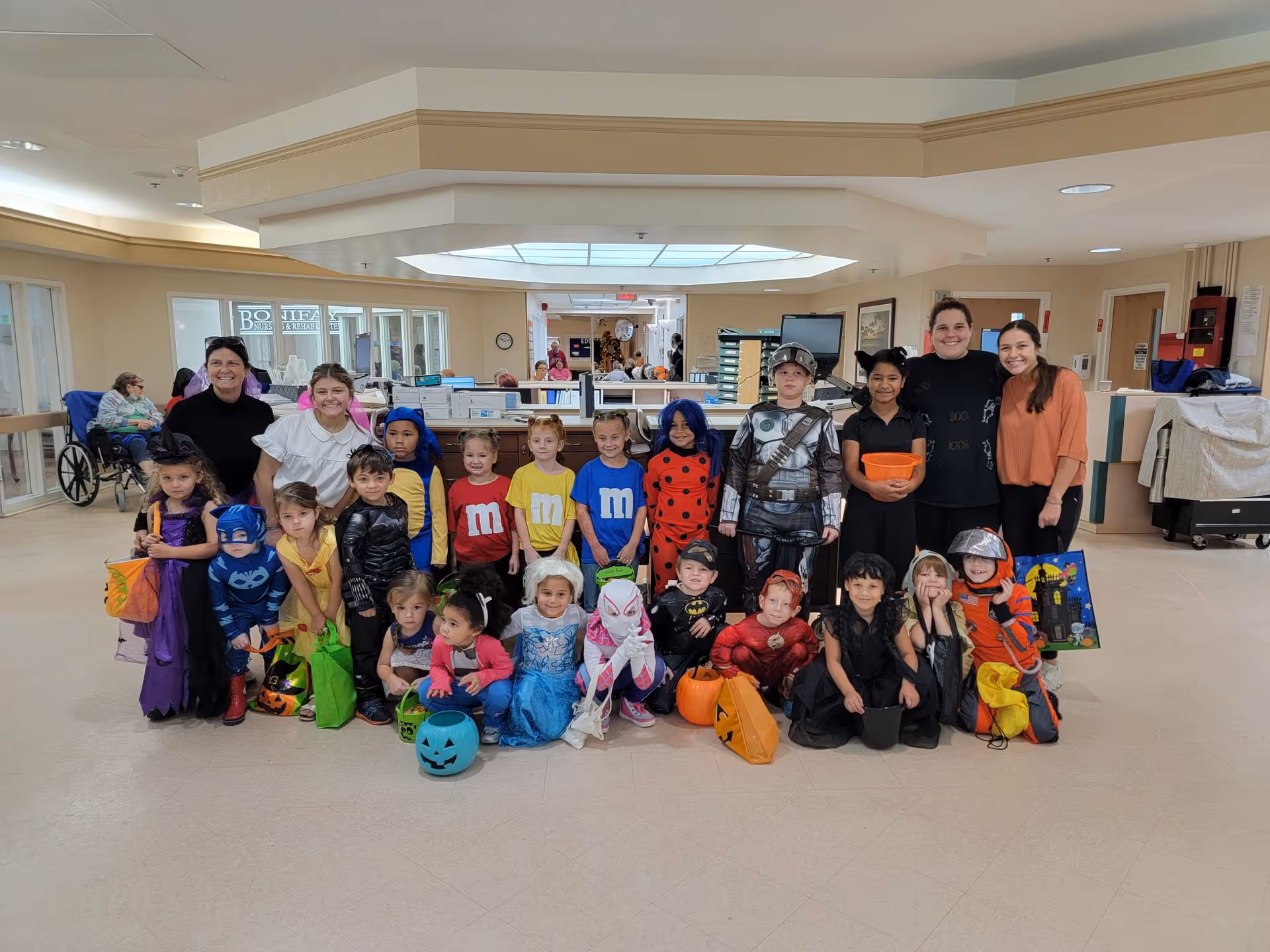 A group of children dressed in various Halloween costumes posing together inside a nursing and rehab center lobby area. Behind them are three adults smiling. The background shows a reception desk and some elderly people sitting in wheelchairs.