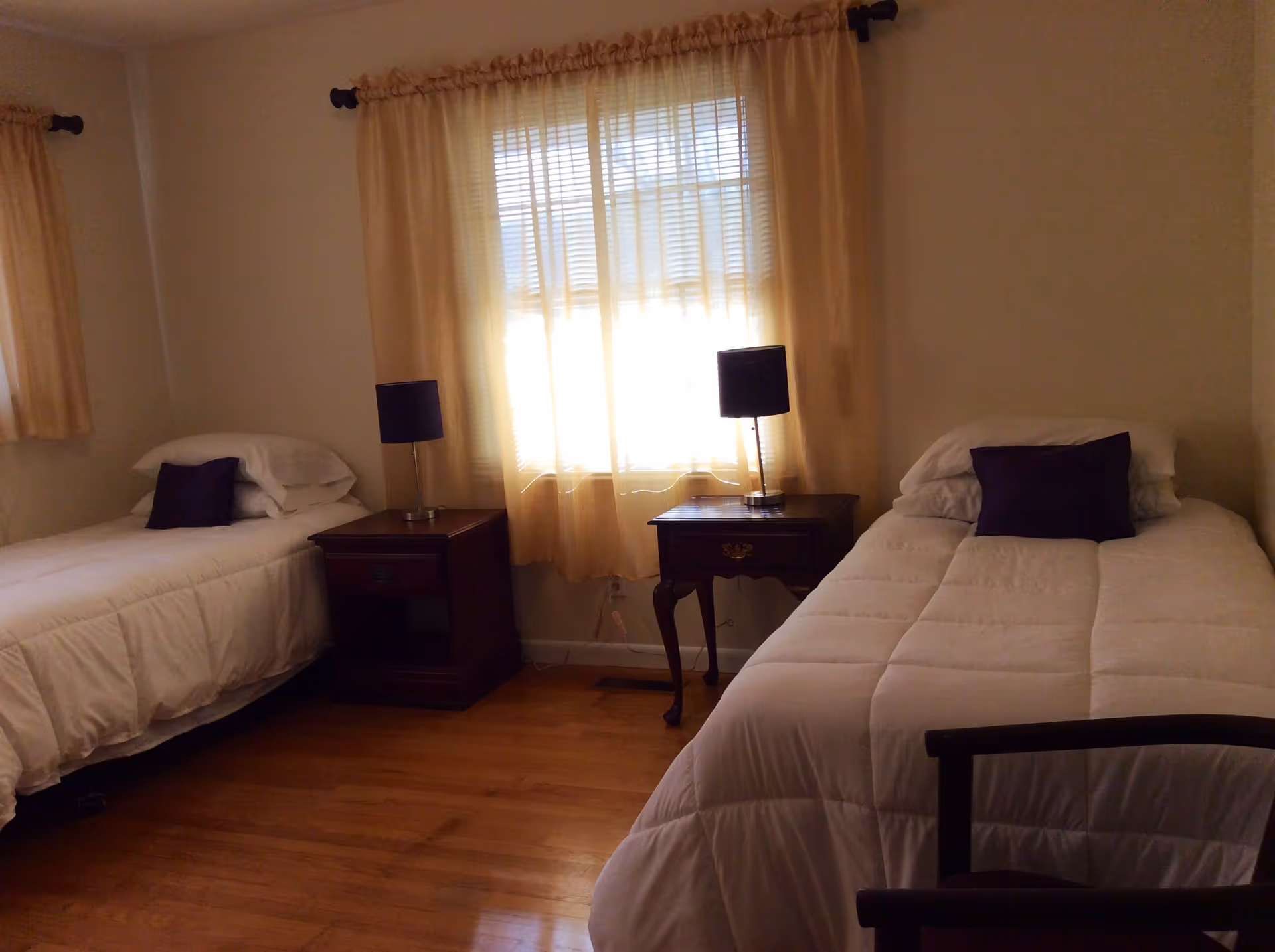 A bedroom with two single beds, each with white bedding and a dark pillow. Between the beds are two wooden nightstands, each with a dark-colored lamp. The room has light-colored walls and wooden flooring, with a window covered by sheer beige curtains letting in natural light.